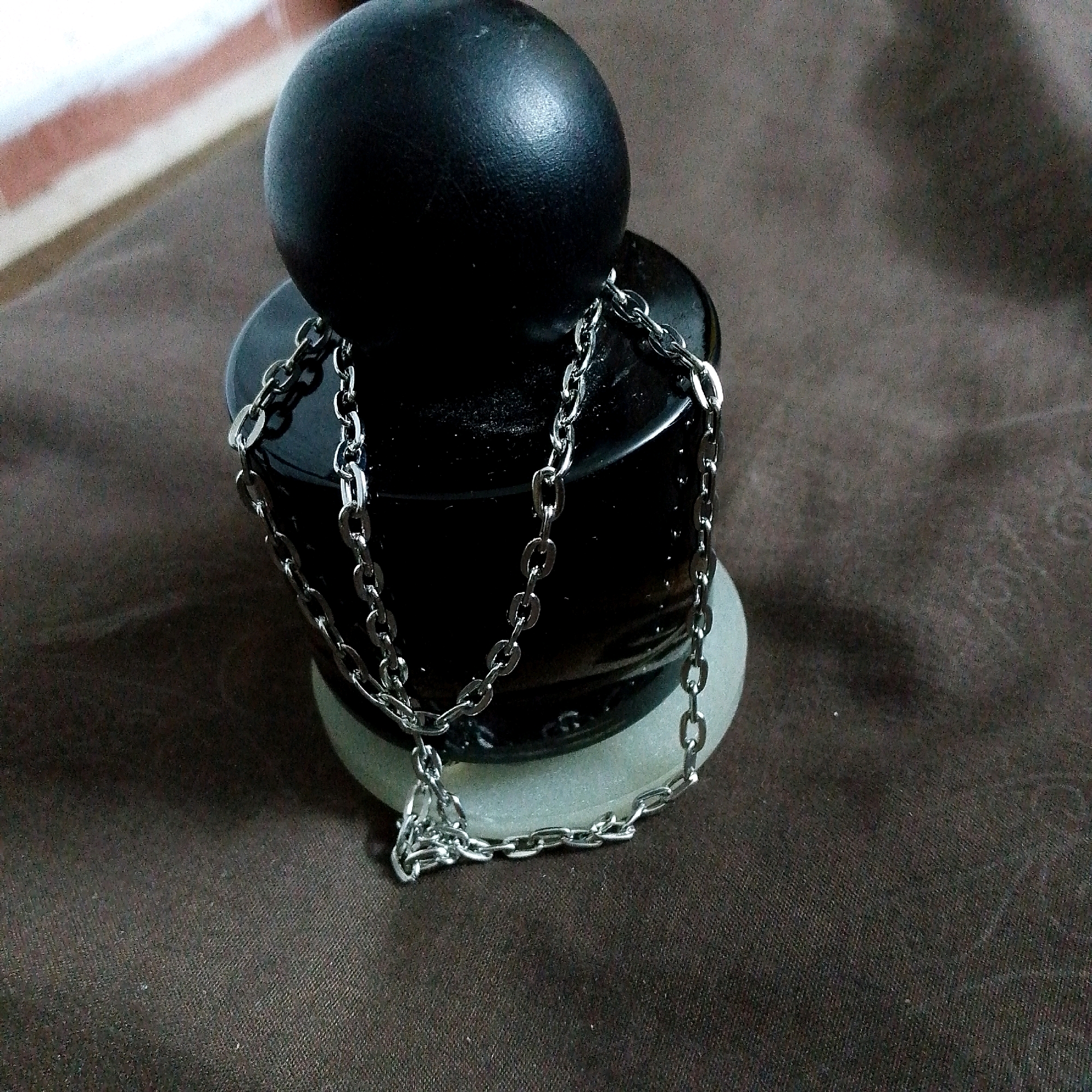 The image shows a close-up view of a black ball with a silver chain draped around it, resting on a dark, textured surface. The ball appears to be made of a matte material, possibly leather or rubber, and has a slightly reflective surface. The chain is made of interconnected oval links. The background is out of focus and appears to be a patterned fabric. There is no discernible location in Irbid, Jordan, or any indication of people, actions, or interactions. The scene is static and focused solely on the object.
