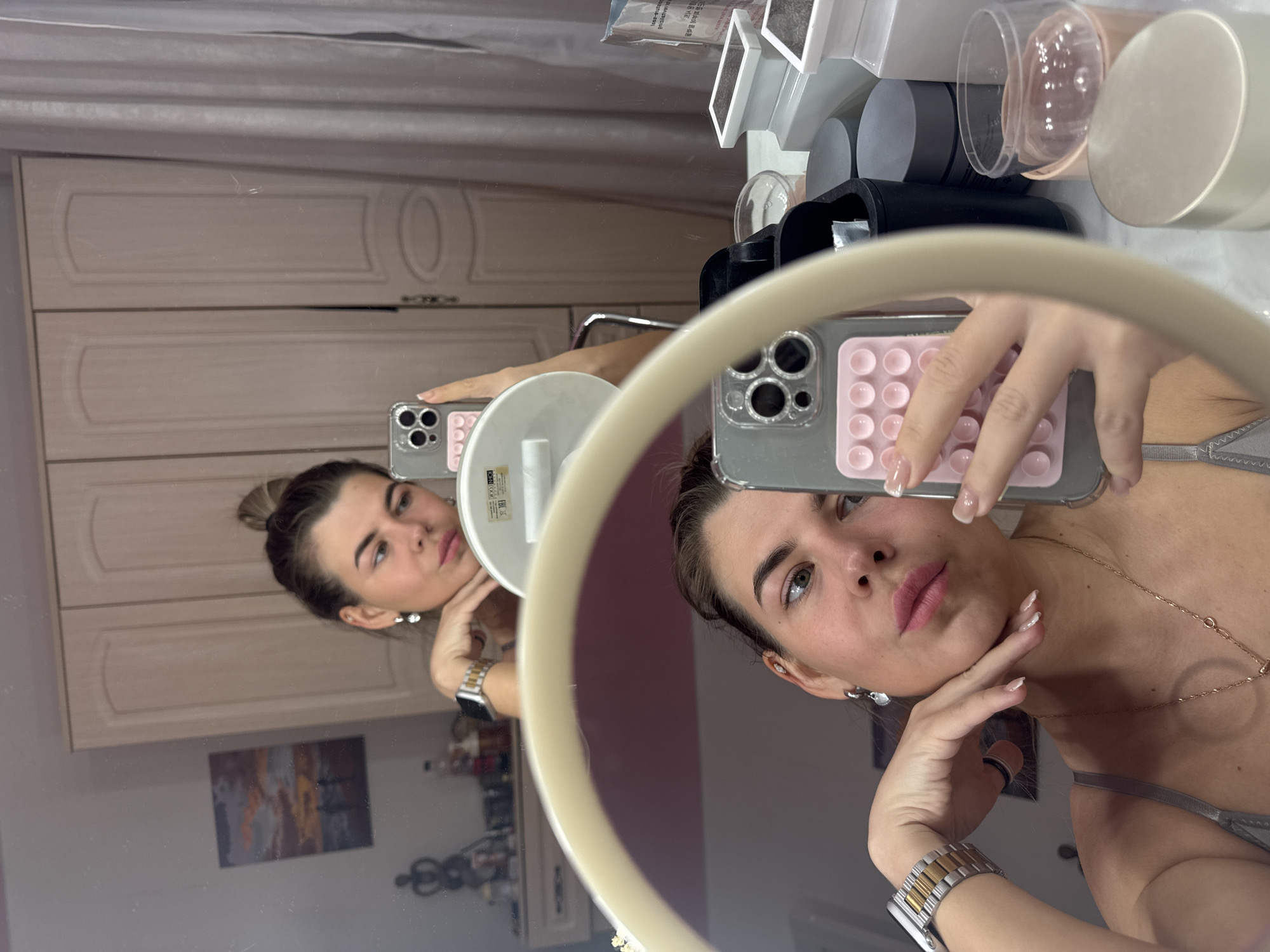 A woman is taking a selfie in a circular mirror. The reflection in the mirror shows her from a different angle, creating a doubled perspective. She is holding a smartphone with a phone case in front of her face. The background includes a vanity with makeup and beauty products on top, and a closet with paneled doors is visible behind the vanity. The location is not discernible as the image does not reveal distinct characteristics related to Chaikovsky, Russia.
