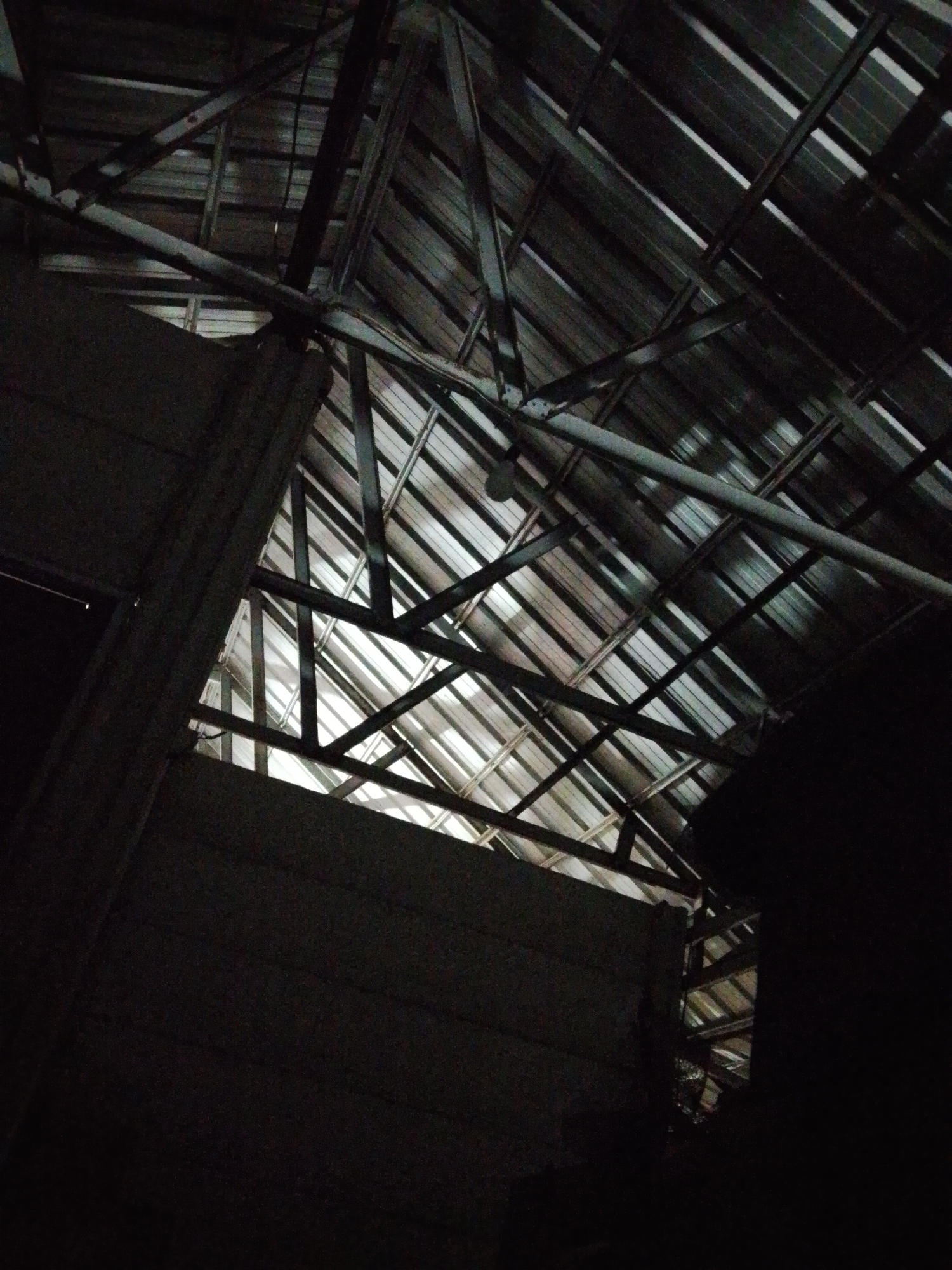The image depicts a dimly lit interior, possibly in Bukit Ketok, Indonesia, revealing the underside of a corrugated metal roof supported by a network of metal beams. A partial wall is visible on the left side, and a dark shape is present on the right side of the frame, obscuring the view. There is a visible gap in the wall, possibly a window or doorway, through which light filters in. The scene appears to be within a structure with minimal ambient light.