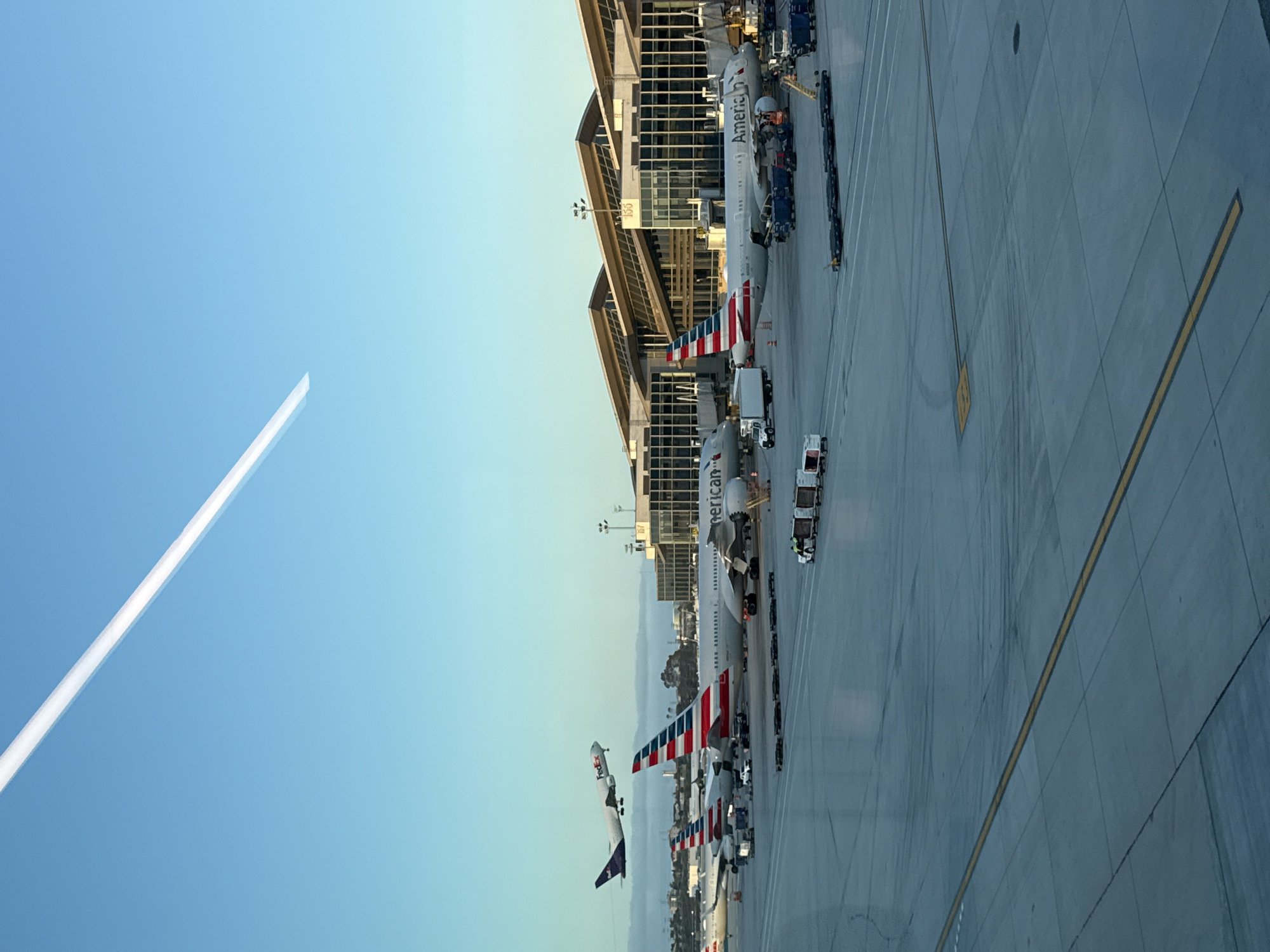 The image depicts an aerial view of an airport tarmac, likely in Los Angeles, California, United States, given the multiple American Airlines planes present. Several American Airlines jets are parked at terminal gates, aligned along the edge of the airport building. Ground personnel and baggage handling equipment are visible around the aircraft. A contrail stretches across the clear, blue sky. The lighting suggests either early morning or late afternoon. A yellow line is on the tarmac.