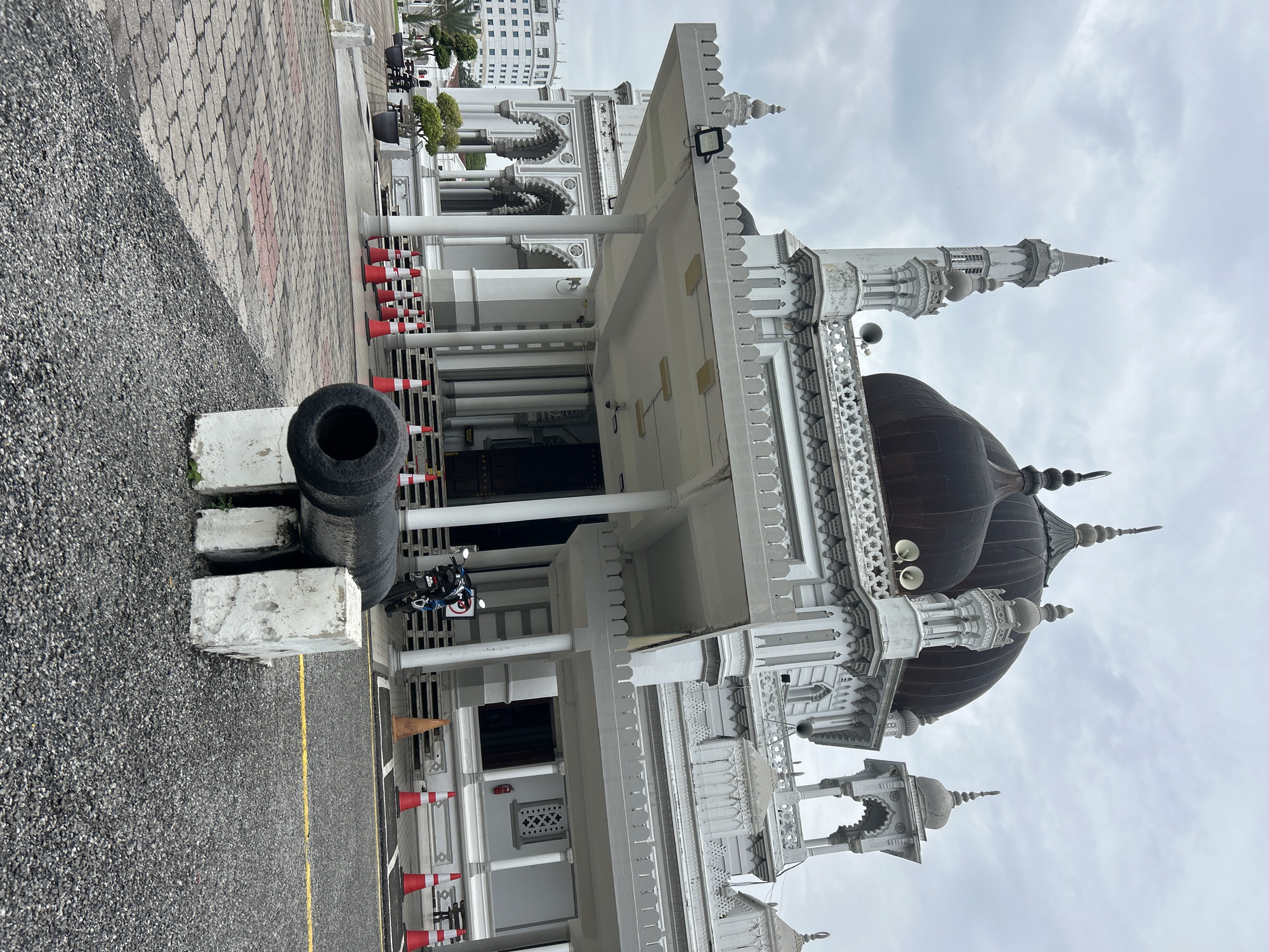 The image depicts the Masjid Zahir in Alor Setar, Malaysia, on an overcast day. In the foreground, a weathered, dark-colored cannon barrel is mounted on two white concrete blocks, positioned on a gravel surface. Behind the cannon, a paved walkway with a patterned design leads towards the mosque's entrance. Red and white traffic cones are lined up along the edge of the walkway. The mosque itself is a grand white structure with intricate architectural details, featuring multiple domes, minarets, and arched doorways. A motorcycle is parked on the steps leading to the entrance. In the background, a modern white building is visible to the left, suggesting an urban setting. The sky is heavily clouded, casting a subdued light on the scene.