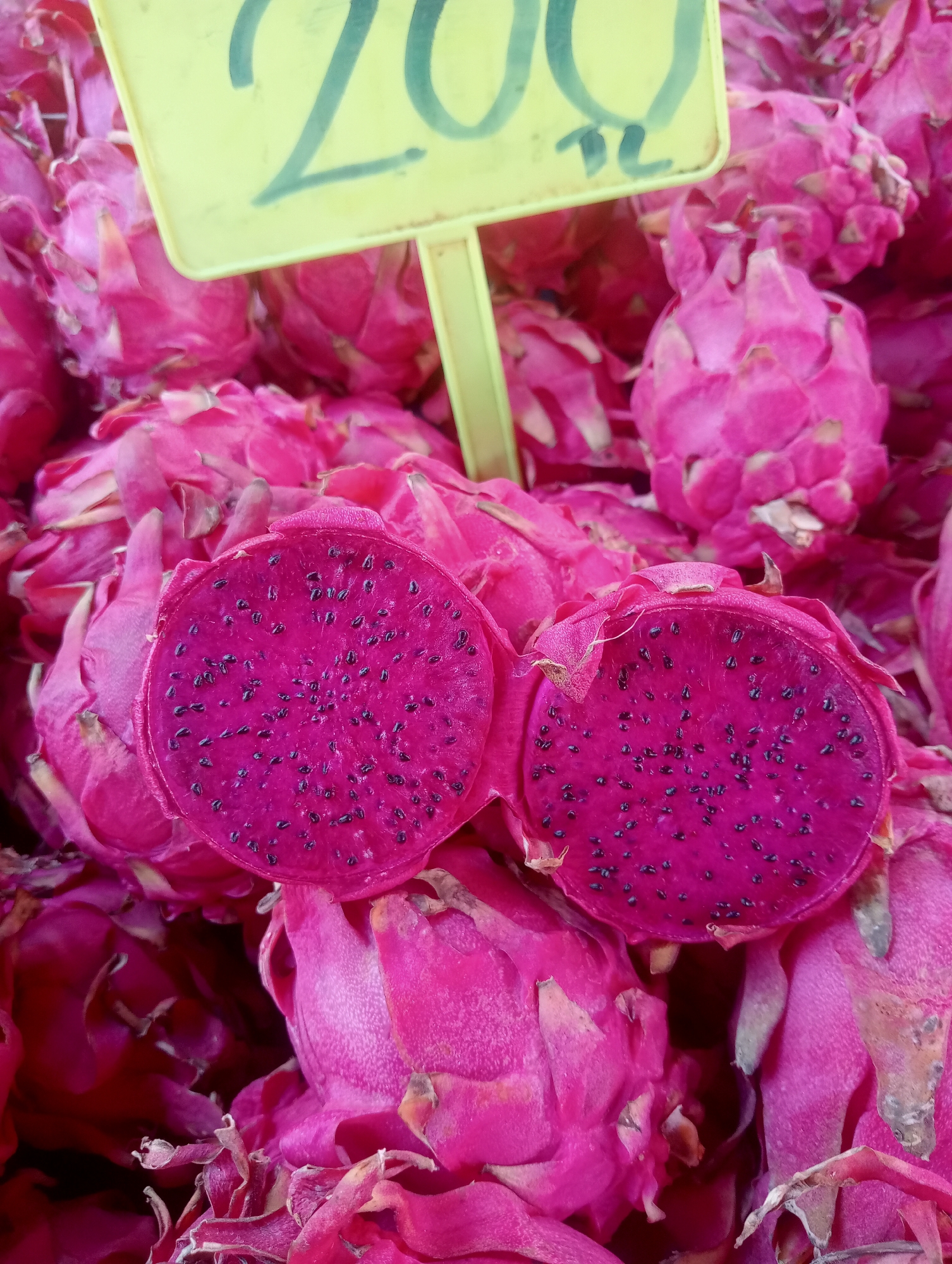 This is a close-up, eye-level, medium shot photograph of a pile of bright pink dragon fruits displayed at a market. A yellow price tag with the number "200" and illegible characters is positioned in the upper left quadrant, partially obscuring the fruit. Two halves of a sliced dragon fruit, revealing its vibrant magenta flesh speckled with numerous black seeds, are prominently featured in the foreground. The fruits appear fresh and are densely packed. The background is filled with more whole dragon fruits. The scene suggests a vibrant, bustling market environment. The image does not provide any specific visual cues to determine its location within the city of Antalya, Turkey.