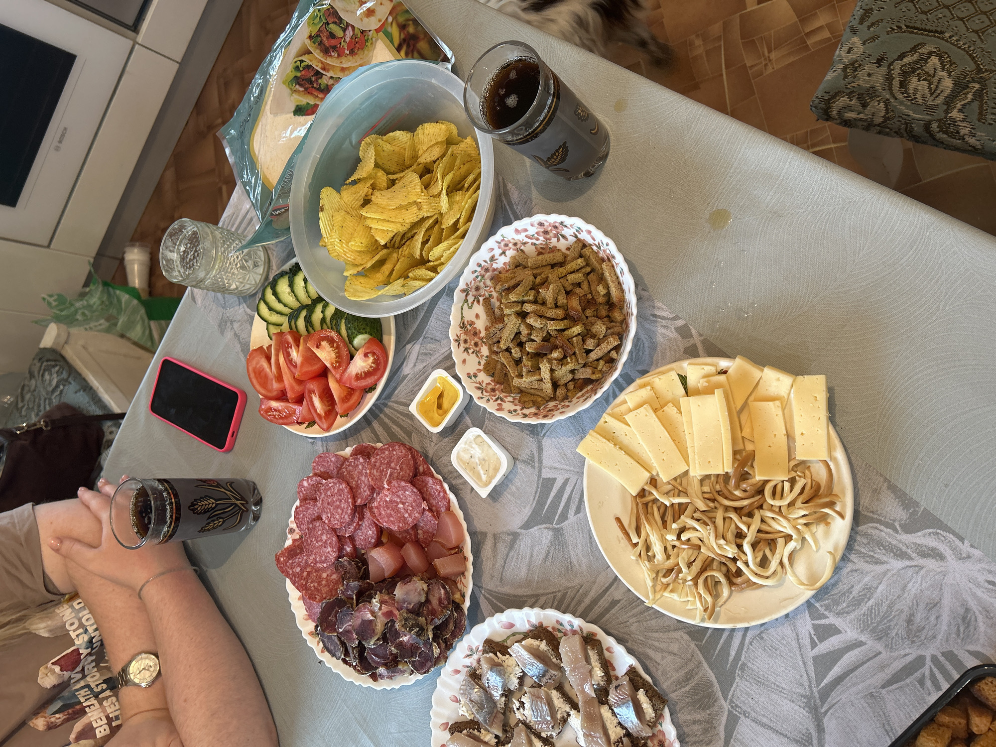 The image shows a table laden with food, likely indoors, in what appears to be a home setting. The dishes include chips, sliced tomatoes and cucumbers, croutons, an assortment of cured meats, cheese slices alongside stringy cheese, and open-faced sandwiches with fish. Condiments, like mustard and a white sauce, are also present. A glass of dark liquid, potentially soda or beer, is visible near the edge of the table next to a hand wearing a wristwatch. There is a mobile phone present, also. A package of food is visible at the top-left. The table is covered with a patterned tablecloth. Based on the information provided, the location is the city of Фоки, Russia.