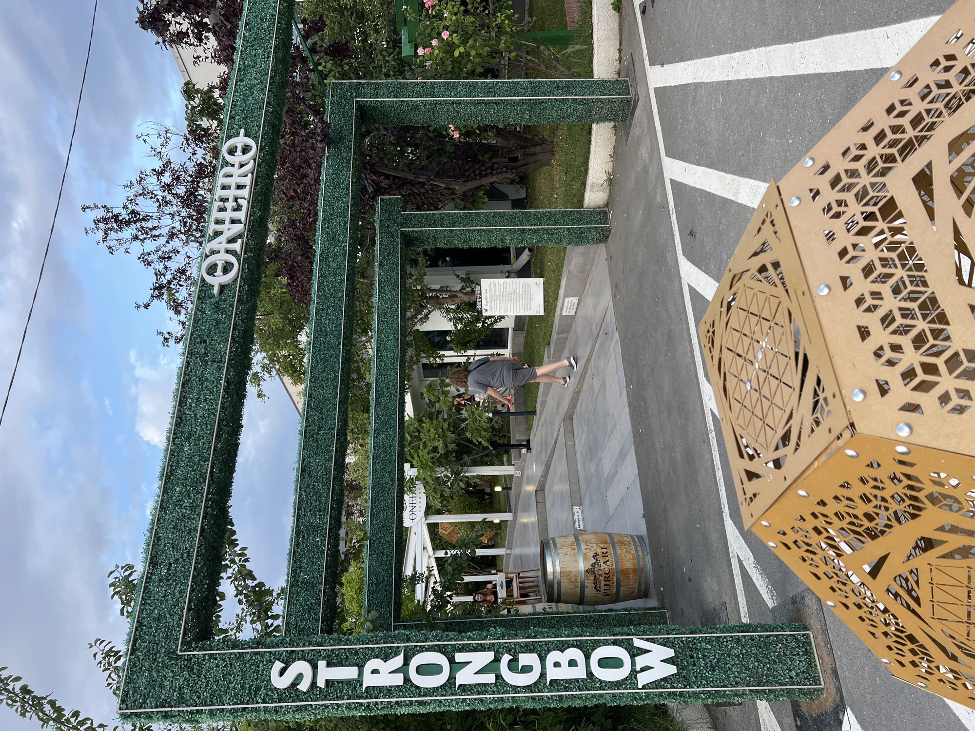 In Năvodari, Romania, a wide-angle shot captures the entrance to a venue, identified by the prominent white lettering "STRONGBOW" vertically on the left and "ONEIRO" horizontally on the right, both set against an artificial green hedge structure that frames an entryway. The entryway leads to a paved walkway, lined with trees and shrubs, with a building visible in the background. A person, dressed in a grey t-shirt and shorts, walks away from the camera towards the building. To the left of the walkway, a wooden barrel with the label "PURCARI" is visible. In the foreground, a geometric, rust-colored metal structure with intricate cutouts sits on a paved surface marked with white lines. The sky above is cloudy with patches of blue.