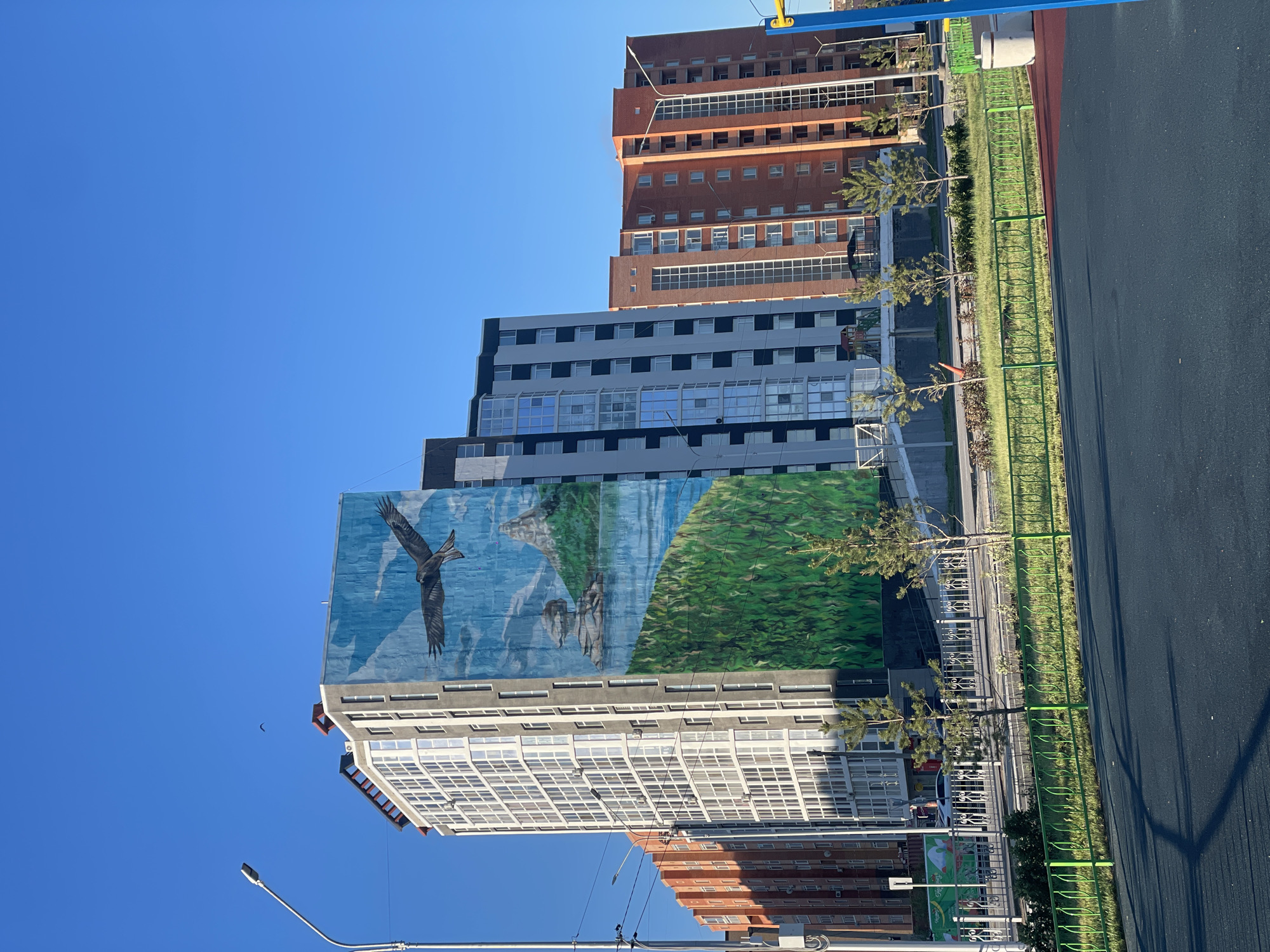 The image shows a view of several multi-story buildings in Kokshetau, Kazakhstan, under a clear blue sky. The buildings have varying facades, including orange, brown, gray, and white sections with numerous windows. The prominent building in the foreground features a large mural depicting a landscape with a soaring eagle. A paved area and green fence line the lower right side of the image. A lamppost and power lines are visible in the lower left corner. There are small trees along the sidewalk.