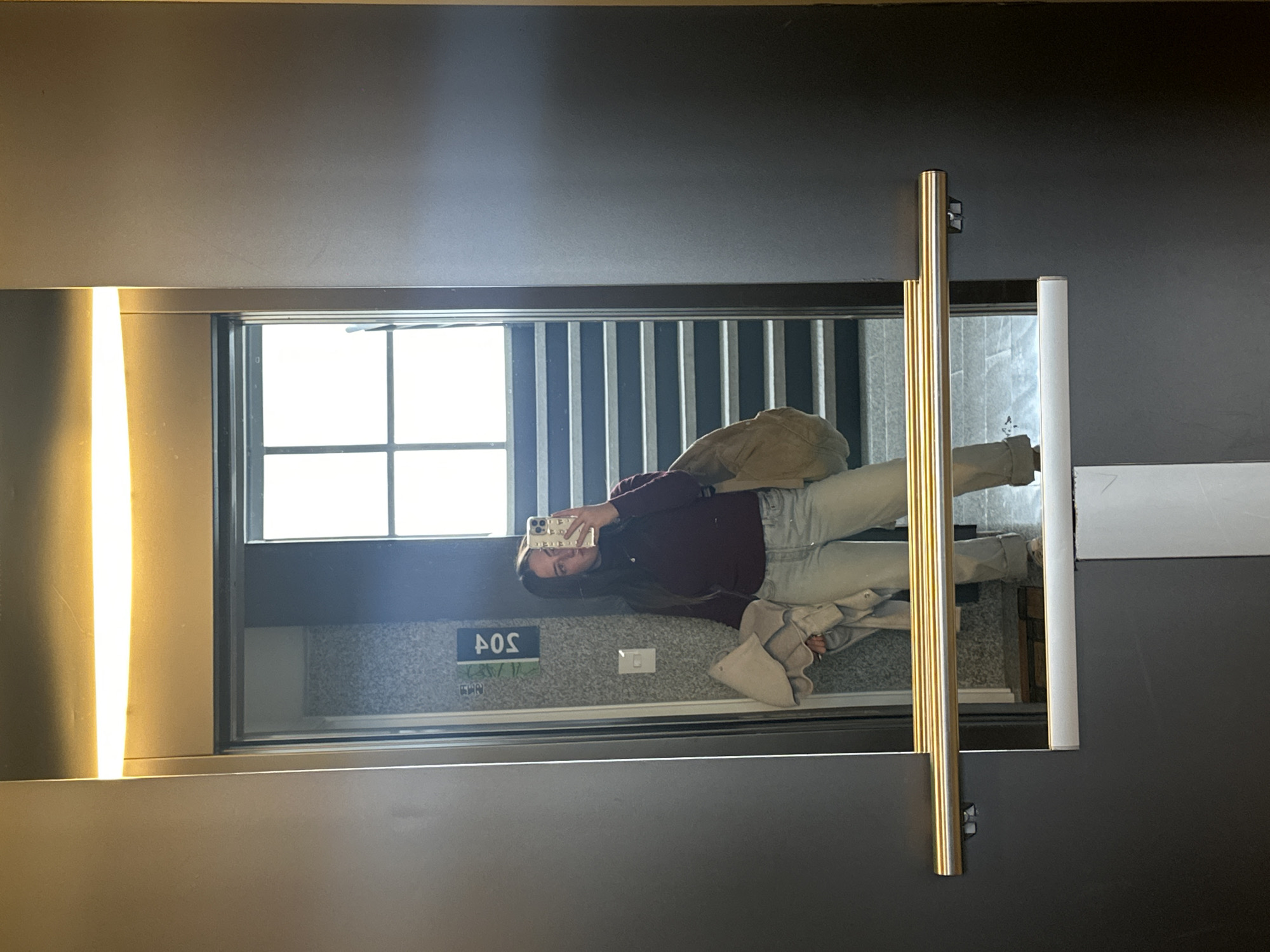 The image, rotated 90 degrees counter-clockwise, depicts a young woman taking a mirror selfie in what appears to be an indoor hallway or common area. She has long, dark hair and is wearing a dark reddish-brown long-sleeved top and light-wash jeans. A light-colored jacket or sweater is draped over her left arm. She holds a white smartphone with both hands, using its camera to capture her reflection in the large, rectangular mirror. Her expression is neutral as she looks towards the phone screen.
The mirror is framed by dark gray or black walls. A bright, vertical strip of light illuminates the scene from the left. A gold-toned vertical handrail stands in front of the mirror, partially obscuring the reflection. In the background reflected within the mirror, a large window with vertical bars or blinds allows bright daylight into the space. Below the window, on a textured gray wall, a white electrical switch is visible. To the left of the switch, an upside-down and mirrored label reads "SO4", which, when correctly interpreted, indicates "405", likely a room number. Smaller, indistinct text is also present below this number. The setting suggests an indoor environment like a hotel, apartment building, or office in Huixquilucan de Degollado, Mexico, during daytime hours.
