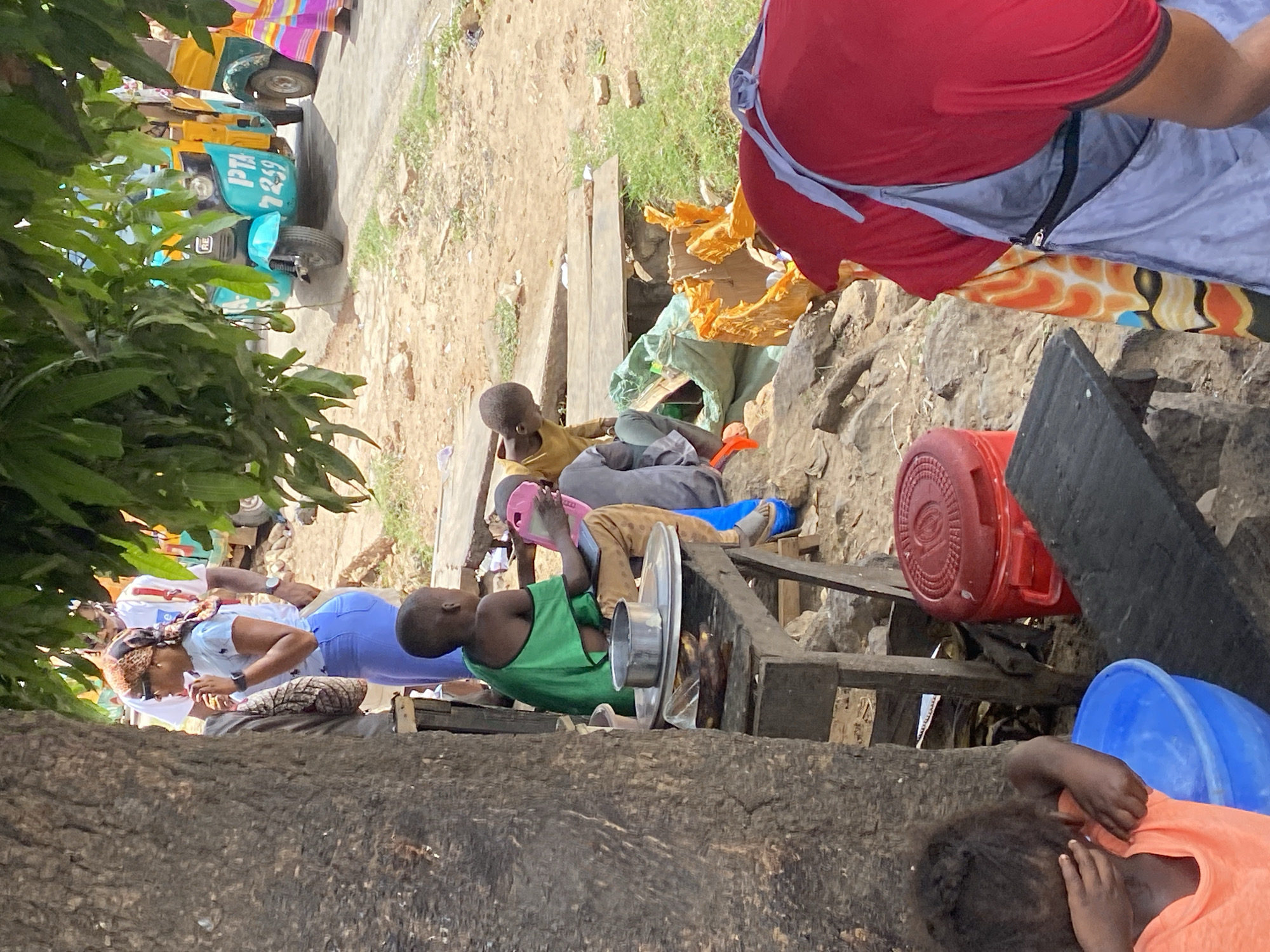 The image depicts an outdoor scene, likely in Jos, Nigeria, based on the presence of a "PTA 7259" marked vehicle typical of that region. A variety of elements are visible: several individuals are present. One woman is seen wearing a white shirt, blue jeans, and a patterned headscarf. Additionally, three children are visible; two are lying down, and one is sitting up. Various items and objects are scattered around the ground, including containers (red and blue), kitchenware (such as a large metal dish), and wooden planks. The environment includes trees, grass, and dirt. Clothing items are spread out on the ground. The overall scene appears to be an everyday street scene with people resting, and objects scattered about.