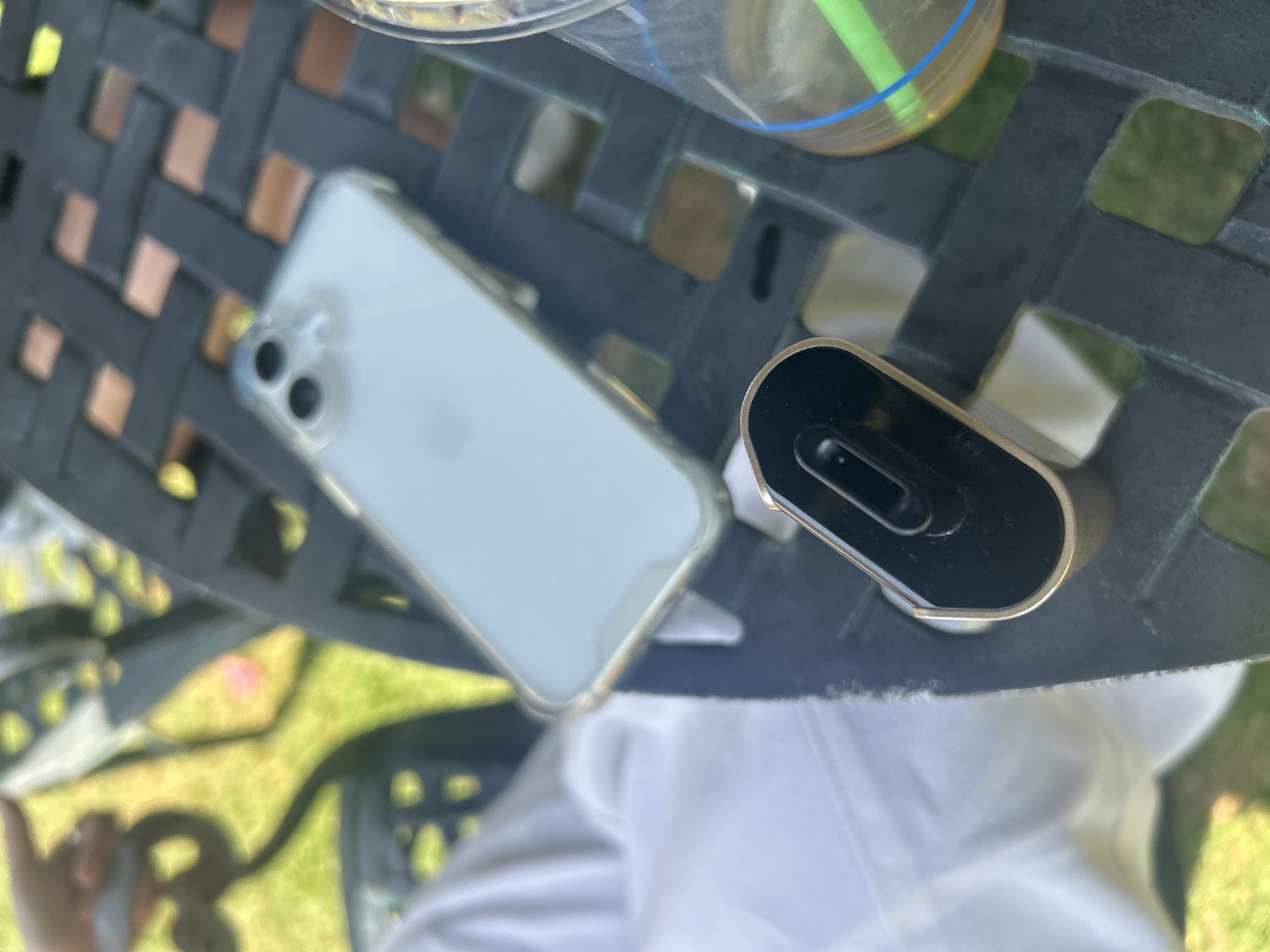 The image shows a table, likely outdoor furniture, with a woven or lattice-like surface. On the table are an iPhone in a clear case, a black and gold object, and a partially visible beverage in a clear cup with a green straw. The background shows blurred green foliage, possibly a lawn or garden, and a glimpse of a white garment, possibly clothing. The objects are angled, suggesting the photo was taken from an elevated or tilted position. There is no information to indicate this is San Bernardino Tlaxcalancingo, Mexico.