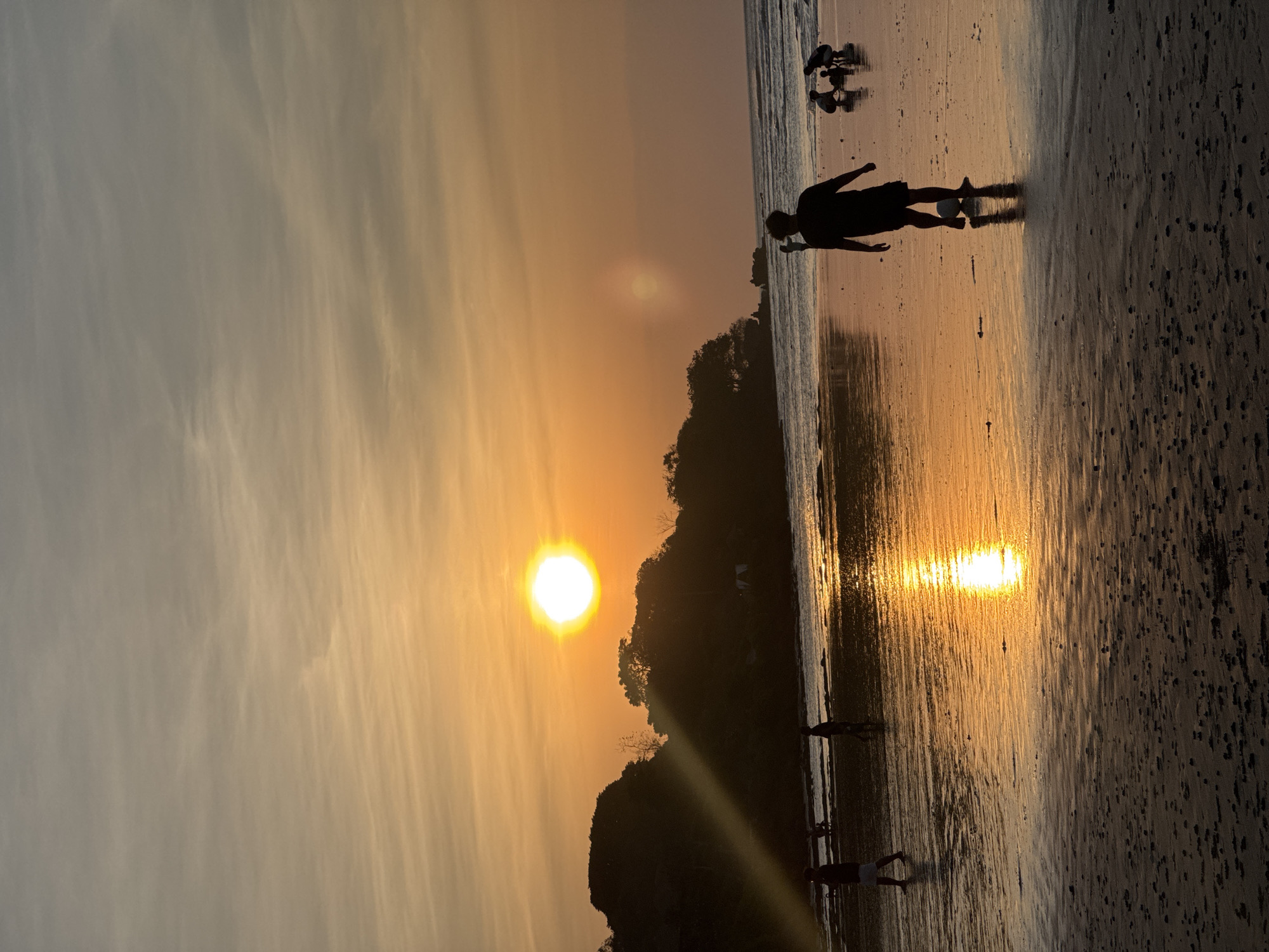 A vertical, silhouette photograph captures the sunset over a beach in Kuta Selatan, Indonesia. The sun, a bright, circular orb, is positioned in the upper right quadrant of the sky, casting a warm, orange glow that illuminates the clouds. A ray of sunlight beams down from the sun, passing through a thick cloud and hitting a dark, tree-covered hillside on the left side of the image. The beach is wet from the receding tide, reflecting the vibrant colors of the sky. In the foreground, a person is silhouetted as they play soccer with a white ball. Further down the beach, other silhouetted figures can be seen, some walking and others gathered together. The ocean is visible in the background, with gentle waves rolling onto the shore. The overall scene is serene and picturesque, highlighting the natural beauty of Kuta Selatan at sunset.
