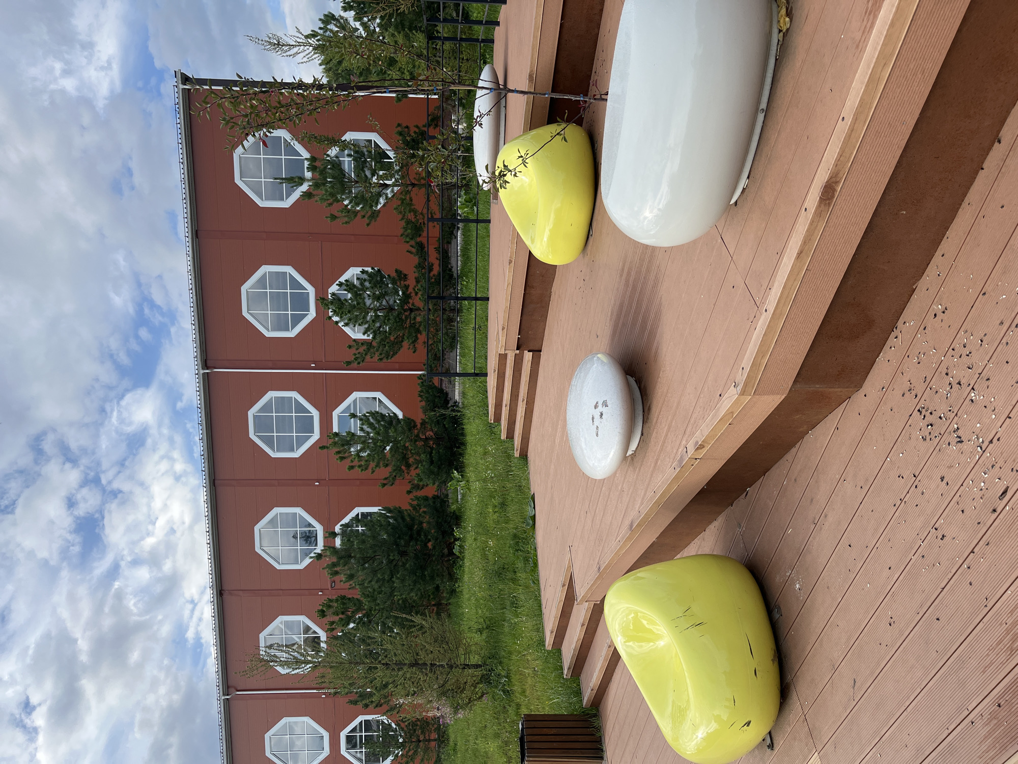 The image depicts an outdoor seating area in front of a modern building with a distinctive red facade and octagonal windows. The building is situated in a green, grassy area with young trees. In the foreground, a wooden deck is furnished with bright yellow and white beanbag-style chairs and two white, smooth, rounded seating structures. There are also steps leading up to the deck, and a trash receptacle on the left side. The sky above is partly cloudy. Based on the architectural style and the context provided, this scene is located in the city of Blagoveshchensk, Russia.