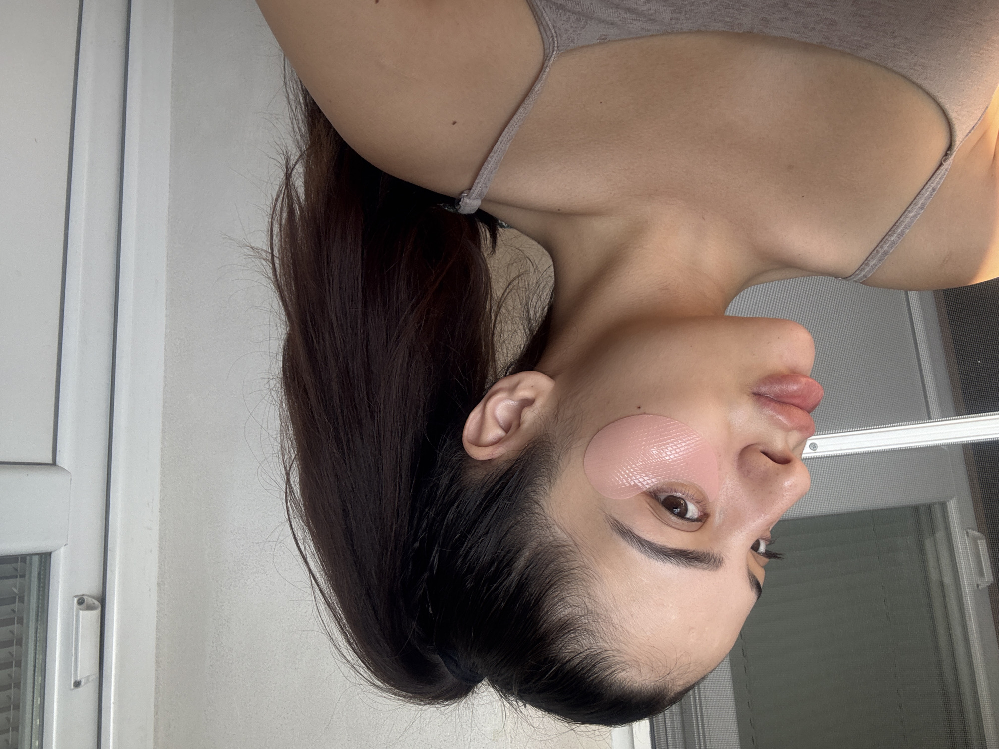 An upside-down portrait-style image, rotated 90 degrees counter-clockwise, depicts a person, likely female, engaged in a self-care routine in an indoor setting in Plovdiv, Bulgaria. The subject has long, dark hair pulled back, revealing her profile. She is wearing a light-colored, possibly grey, tank top or camisole.
The notable activity is the application of two pink, textured eye patches beneath her left eye. Her gaze is directed towards the right side of the frame, beyond the camera, and her lips are slightly parted, conveying a neutral or focused expression.
The background consists of a plain, light-colored wall. To the left (which would be the bottom if the image were correctly oriented), a white-framed window is partially visible, featuring a screen or blinds behind its glass panes. The scene is illuminated by natural, bright daylight, suggesting it is daytime, with a soft, warm highlight on her neck and right shoulder from the light source. No visible text is present in the image.