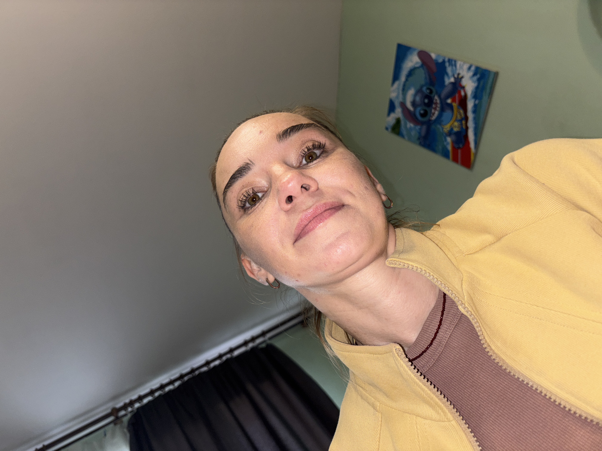 The image shows a low-angle, close-up, selfie-style shot of a young Caucasian woman. She is wearing a yellow zip-up jacket over a mauve ribbed t-shirt. Her hair is pulled back, and she is looking up at the camera. She has light brown eyes, visible eyelashes and eyebrows, and light pink lipstick. A subtle double chin is visible. A small, dark bead necklace is worn around her neck. In the background, a dark curtain or drape is visible on the left, and a light green wall with a framed picture of the character Stitch from Disney's Lilo & Stitch is on the right. The overall environment appears to be indoors, possibly a bedroom. There is no discernible indication of the image being located in Sinyavino, Russia.