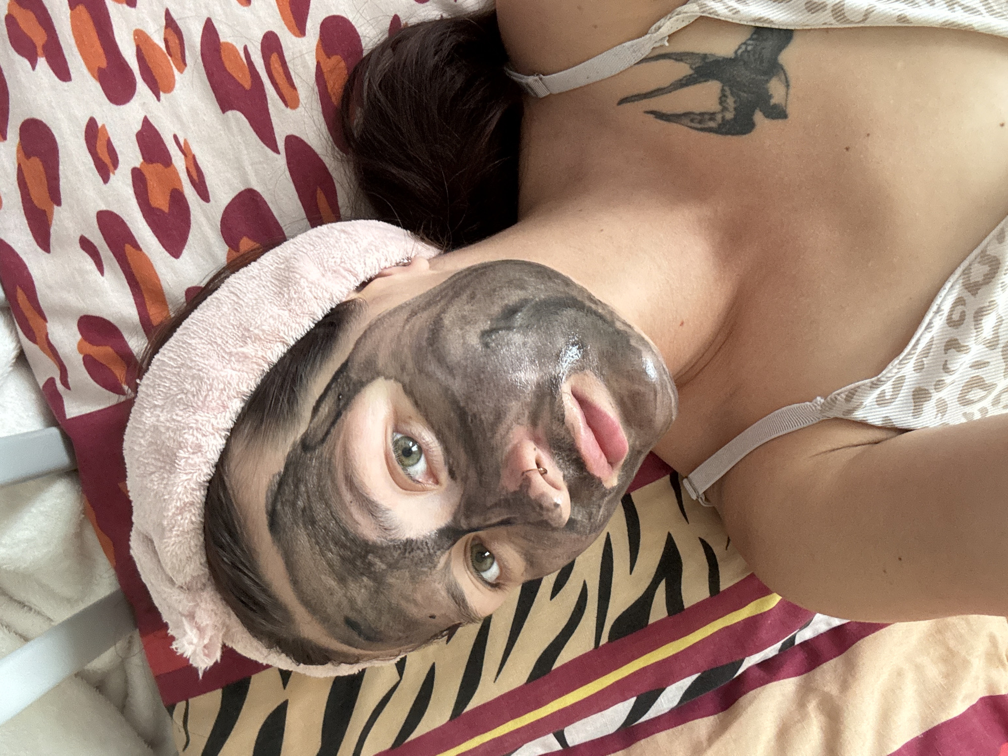 A close-up, overhead shot shows a young Caucasian woman lying down with a black charcoal face mask applied. She is wearing a pink terrycloth headband. Her eyes are green and open, and she has a nose ring. A tattoo of a swallow is visible on her upper chest, just below her right collarbone. She is wearing a light-colored camisole with a leopard print. The background consists of patterned bedding, featuring stripes of maroon, yellow, and beige, and a leopard print. The scene appears to be in a bedroom. No specific location in Naberezhnye Chelny, Russia can be determined from the image.