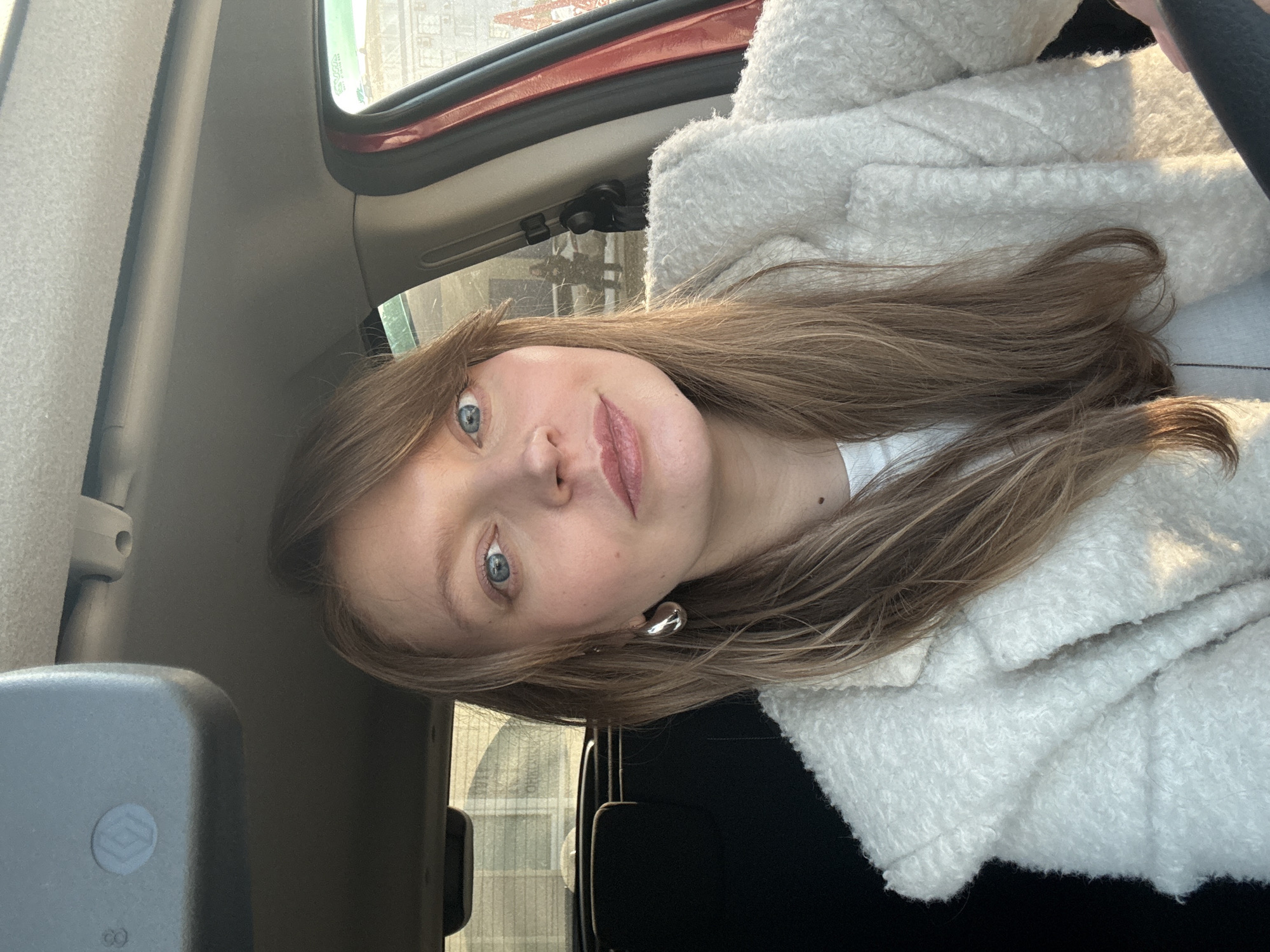The image captures a female individual inside a vehicle, located in Izhevsk, Russia. The subject, a Caucasian female with light brown hair parted on the side and blue eyes, is positioned in what appears to be the driver's seat. She is wearing a white, textured, long-sleeved outer garment over a white undergarment. A silver, teardrop-shaped earring is present on her left ear. Her gaze is directed towards the camera with a neutral expression.
The vehicle's interior shows a rearview mirror with a diamond-shaped logo (Renault) in the upper center, a light-colored ceiling, and a reddish-orange section of the front passenger door frame on the right. A portion of a dark steering wheel is visible in the lower right.
The exterior environment, viewed through the vehicle windows, includes: through the left rear window, a blurry view of a light-colored structure. Through the front passenger window on the right, a green sign with white Cyrillic text 'АВТОМОЙКА 24 ЧАСА' (Car Wash 24 Hours) is visible. Beyond the sign, a light-colored building with red markings and a blurred outline of a person and another vehicle are discernible, indicating a commercial or street setting.