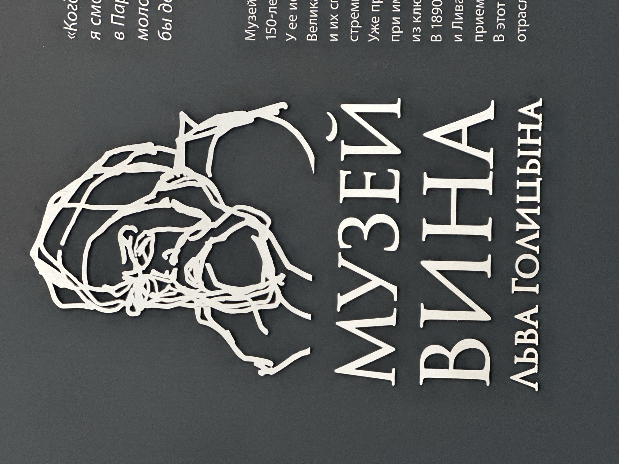 A close-up, eye-level, medium shot shows a dark gray, textured wall with metallic letters and a metallic wireframe outline of a man. The wireframe depicts a man's head in profile facing left, with his face etched with lines, suggesting an older man with a beard and glasses. To the right of the wireframe is a block of text in a light-colored, sans-serif font. The text is in Russian and appears to be a description or quote. Below the wireframe and to the left of the text, large metallic letters spell out "МУЗЕЙ ВИНА ЛЬВА ГОЛИЦЫНА" which translates to "Museum of Wine of Lev Golitsyn". The lighting is even and bright, creating a slight shadow from the wireframe on the wall.