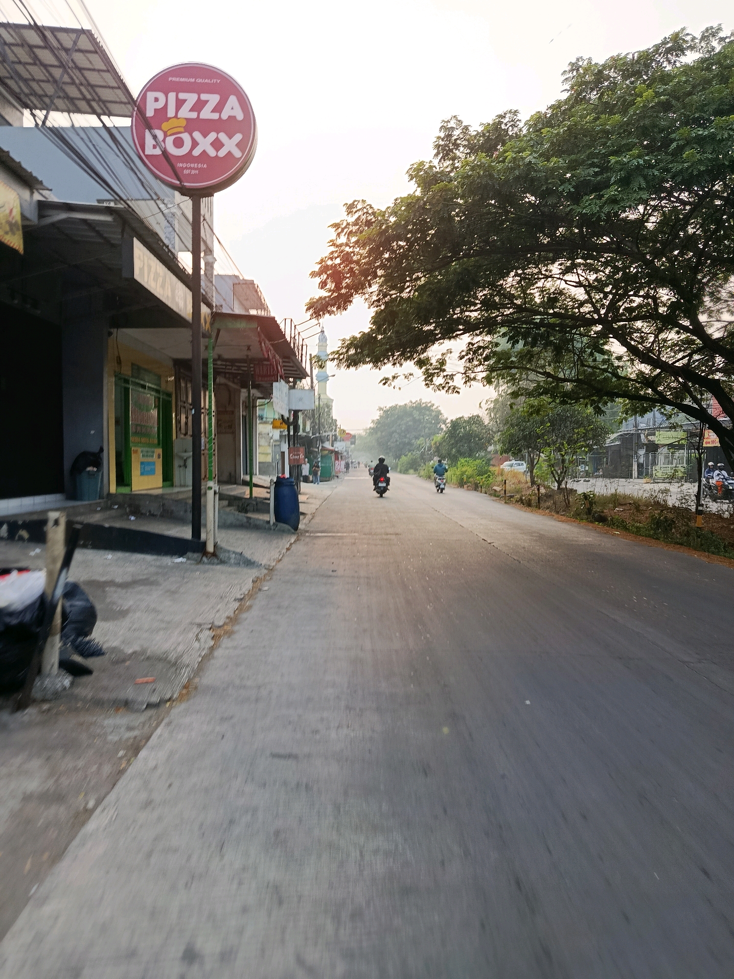 An outdoor, eye-level, vertical shot captures a street scene in Bekasi, Indonesia, during the early morning or late afternoon. The foreground is dominated by a sidewalk and a paved road that recedes into the distance. On the left, a commercial building features a prominent red circular sign with "PIZZA BOXX" in white letters and a yellow chef's hat graphic. Below the sign, a dark gray trash can stands next to a sidewalk. The sidewalk is concrete and shows signs of wear and tear, with some debris visible.
The road is asphalt and appears to be a two-lane street with traffic moving away from the camera. Several individuals on motorcycles are present on the road, with one rider in the middle distance wearing a white helmet and a dark blue shirt, and another further back wearing a black helmet. On the right side of the road, a large tree with dense green foliage extends into the frame, partially obscuring the sky. Beyond the tree, more buildings and vegetation can be seen, and a white mosque minaret is visible in the background. The sky is a pale yellow, suggesting either sunrise or sunset. The overall atmosphere is quiet and suggests a typical urban street in Indonesia.