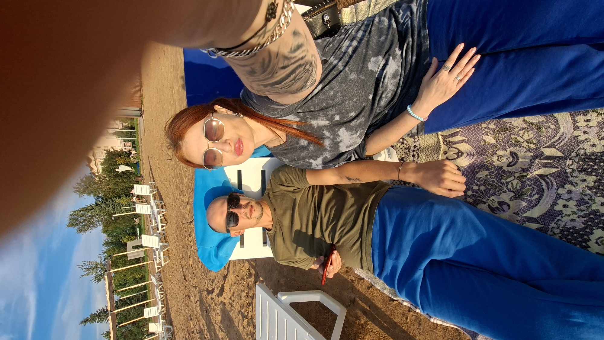 Two people, a man and a woman, are lying on a beach on a sunny day. The man, on the left, is wearing a green t-shirt and blue pants and is holding a red phone. He is wearing sunglasses. The woman, on the right, is wearing a grey tie-dye shirt and blue pants, and is also wearing sunglasses. She has red hair and is wearing jewelry on her wrist. They are lying on lounge chairs on a sandy beach with buildings and trees in the background. This location is presumed to be in the city of Myadzyel, Belarus.