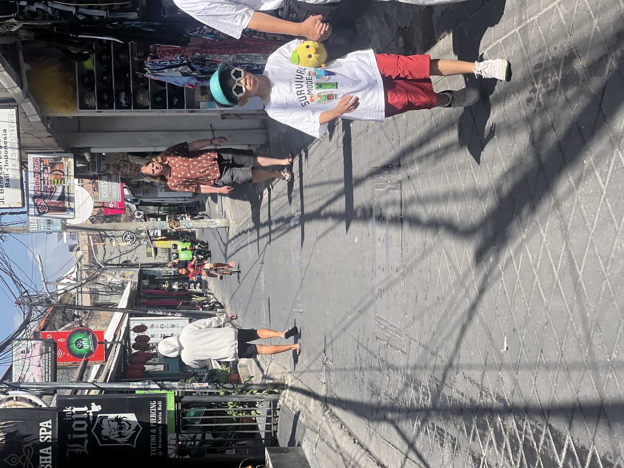 The image depicts a narrow, paved street in Kuta, Bali, Indonesia, lined with shops and adorned with numerous overhead electrical wires. Sunlight casts long shadows across the textured, diamond-patterned pavement. In the foreground, a young boy wearing a white t-shirt with a Minecraft "Survival Mode" graphic, red shorts, white sneakers, and a turquoise baseball cap with star-shaped sunglasses walks forward, holding a yellow plush toy in his right hand. A person's arm, holding the boy's hand, is visible on the right. Behind the boy, a person with long blonde hair, wearing a patterned brown shirt and dark shorts, walks away from the camera. Further down the street, a person in a white hooded sweatshirt and dark shorts walks away from the camera, and a child rides a bicycle. Several shops are visible, including one with a large black sign for "SHA SPA" and another with a red circular sign featuring a green stylized lion. Other signs indicate tattoo and piercing services, leather crafts, and various other businesses. Motorbikes are parked along the sides of the street.
