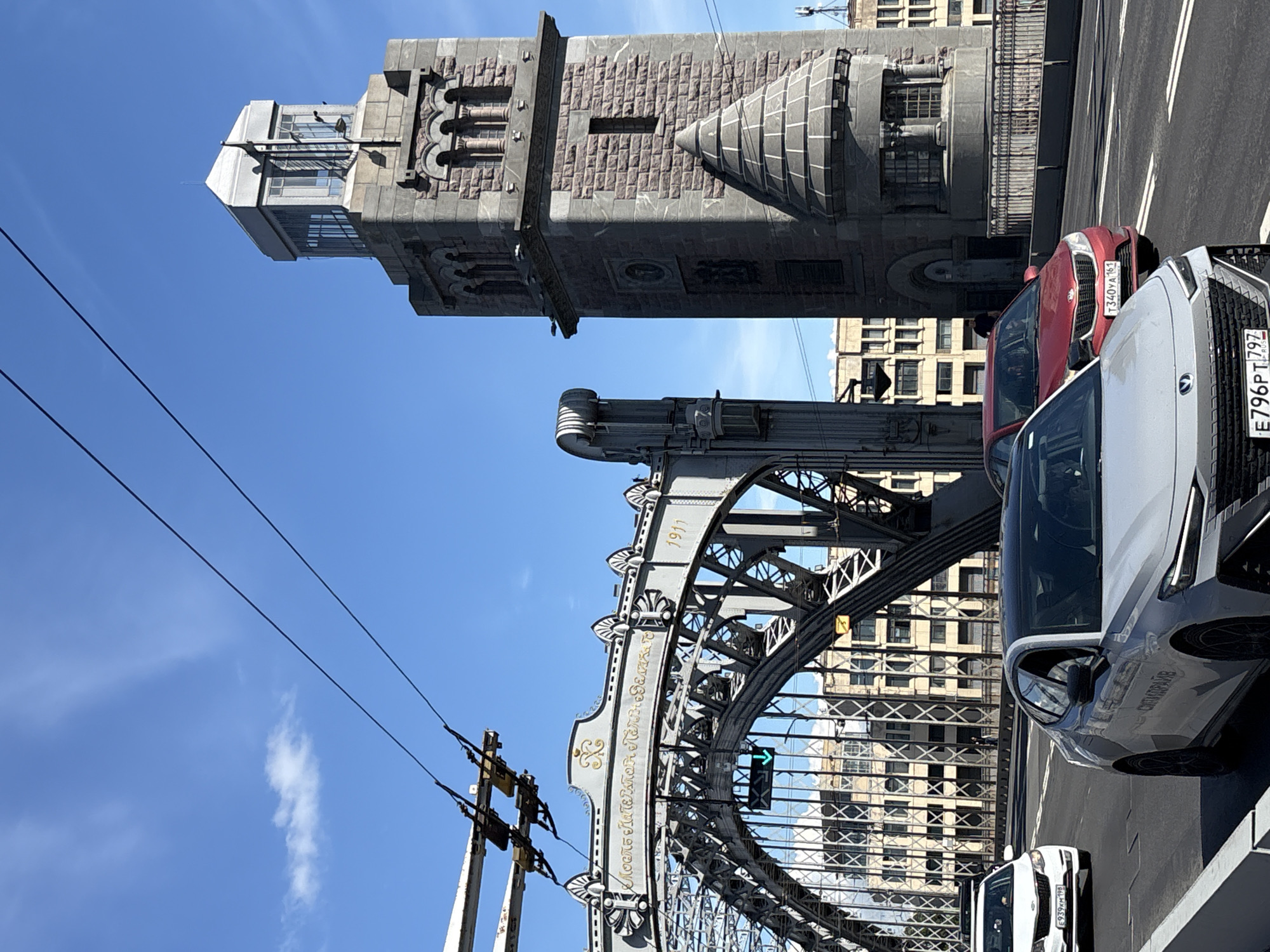 This media file captures a bright, sunny day in Saint Petersburg, Russia, with a clear blue sky and a few wispy clouds. The perspective is looking upwards and slightly to the side, presenting a striking view of the Trinity Bridge and its distinctive towers. The bridge's intricate metalwork, painted a dark gray, is prominently featured, with the year "1911" visible on one of its decorative elements. The bridge is adorned with ornate golden lettering, which appears to read "МОСТ ИМПЕРАТОРА АЛЕКСАНДРА III" (Bridge of Emperor Alexander III).
In the foreground, three cars are parked or driving on the bridge. The closest vehicle is a silver SUV, partially obscuring the view of a white sedan behind it. A red car is visible further back. The license plates of the silver SUV are partially visible, reading "E796PT797" and a partial Russian text. The red car's license plate is "T3340YA163". The background is dominated by a multi-story building with a light-colored facade and numerous windows, reflecting the sunlight. The imposing stone tower of the bridge, with its textured masonry and architectural details, is a significant focal point, suggesting a historically rich location. The scene conveys a sense of urban grandeur and historical significance. The time of day is likely daytime, given the strong sunlight and clear sky.