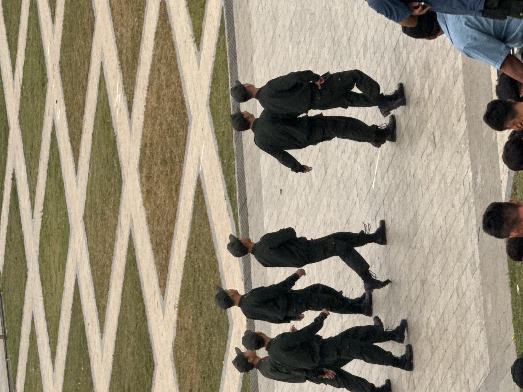 The image shows a group of people dressed in black uniforms and black caps, standing in formation on a paved area. Some of the individuals are in focus and clearly visible, while others are partially obscured or out of focus. The paved area appears to be a walkway or plaza, with a diamond-patterned design in alternating sections of grass and lighter paving stones. The lighting suggests it is daytime, possibly late afternoon, given the slightly muted colors. The people are standing with their backs to the camera, facing towards the right side of the frame. Their postures suggest they are participating in some sort of organized activity, possibly a ceremonial march or formation. No text is visible in the image. The overall impression is one of discipline and order, with the individuals appearing serious and focused on their task. The scene is outdoors, and the environment appears to be a public space, possibly a park or a memorial.