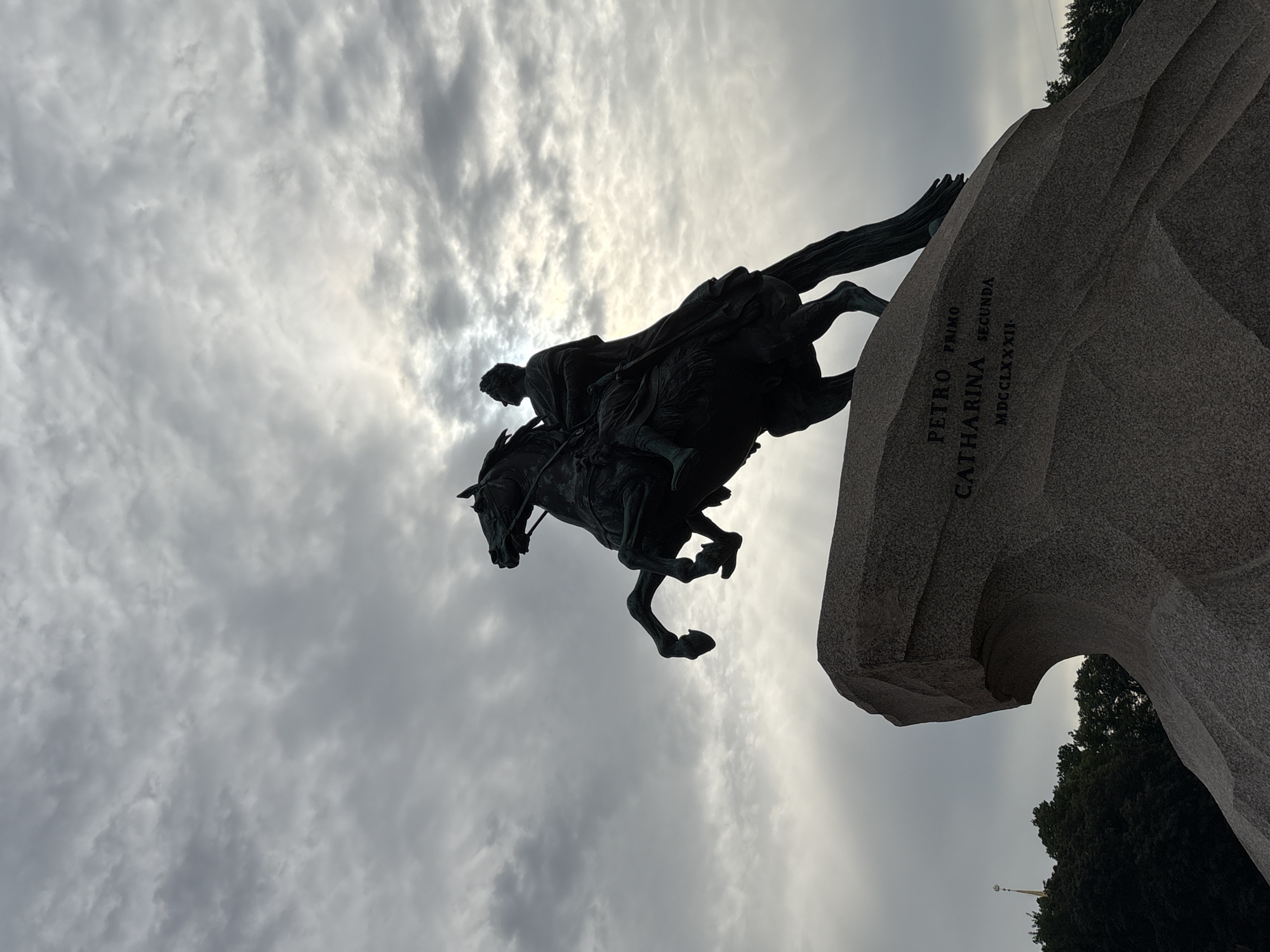This is a medium shot, low-angle, silhouetted photograph of the Bronze Horseman monument in St. Petersburg, Russia. The iconic equestrian statue of Peter the Great, cast in bronze, is depicted rearing on its hind legs atop a large, irregular granite pedestal. The monument is set against a cloudy sky with the sun partially visible behind the clouds, creating a dramatic halo effect. The base of the monument is inscribed with Latin text: "PETRO PRIMO CATHARINA SECUNDA MDCCLXXXII". In the background, dark silhouettes of trees and a distant spire are visible. The photograph captures the monument's imposing scale and its powerful, dynamic pose.