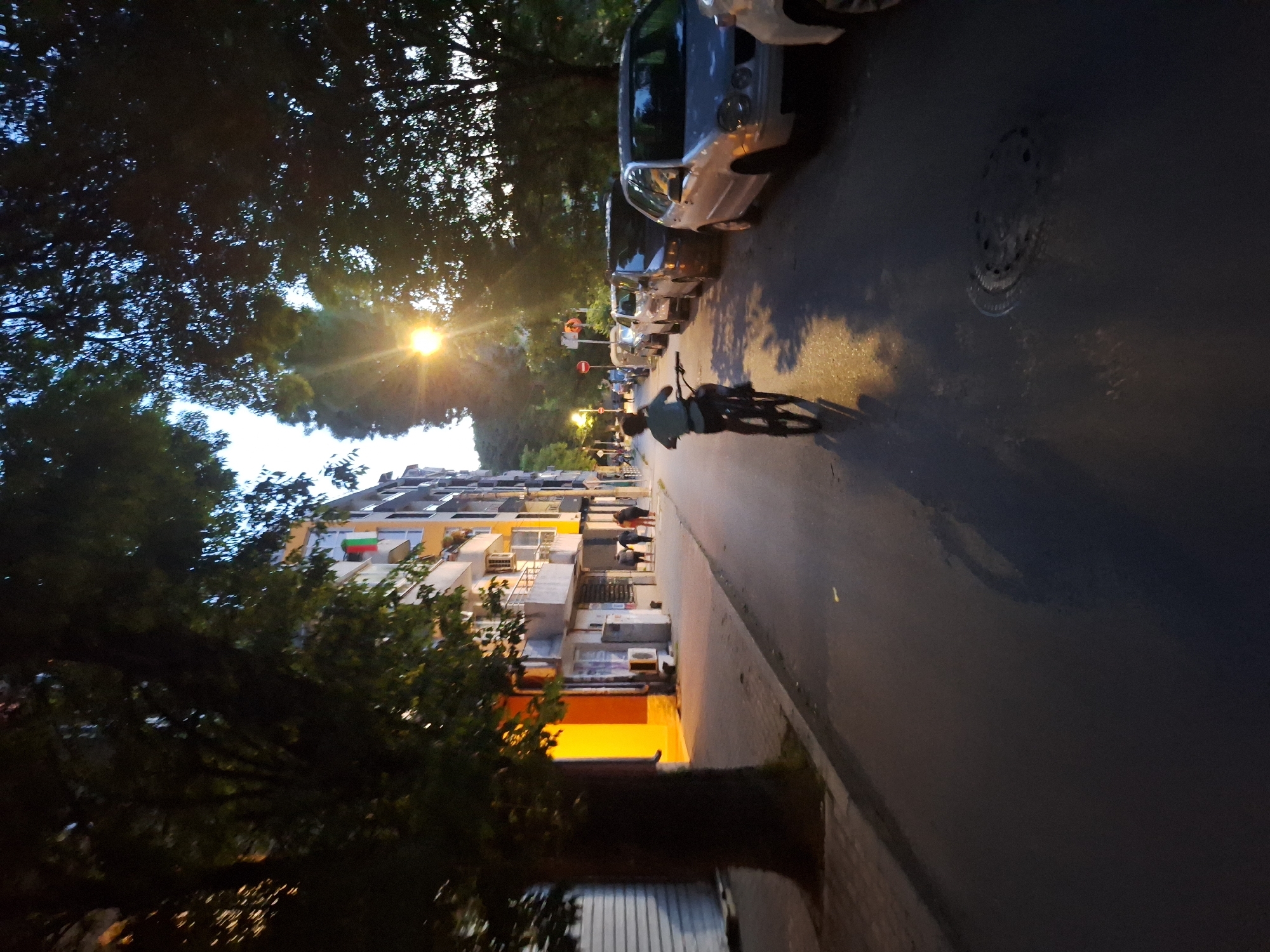 A young person on a bicycle rides down a street in Varna, Bulgaria, during dusk. The street is lined with trees and buildings, and cars are parked on the side. A street lamp illuminates the scene.