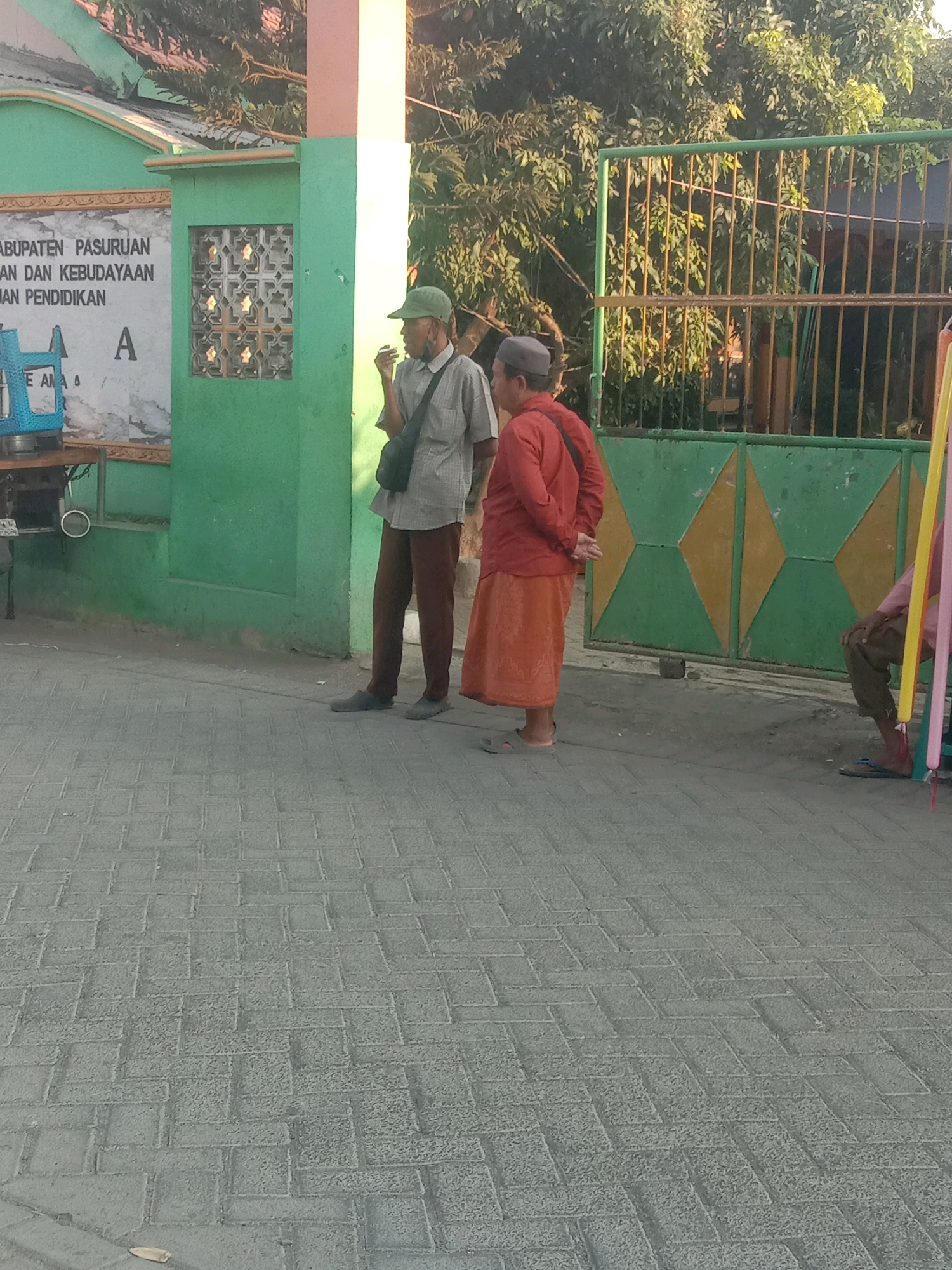 In the city of Selotambak, Indonesia, two men stand near a green gated entrance. The man on the left wears a light-colored, short-sleeved collared shirt, brown pants, a green cap, and a dark shoulder bag. He is holding something to his mouth, possibly a cigarette. The man on the right wears a red long-sleeved shirt and an orange skirt, with a dark shoulder bag. He is facing away from the camera. A portion of a blue dispenser with a white top is visible on the left side of the image, and a green wall with a decorative window is behind the man in the green cap. The ground is paved with gray interlocking bricks. The background features green foliage and a yellow and green patterned gate.
