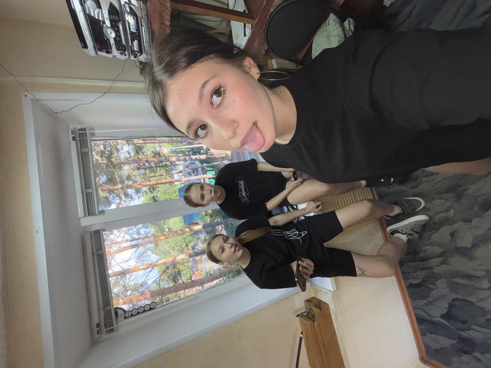 The image captures three young women indoors in Chaikovsky, Russia, during what appears to be a bright daytime. The photograph is a selfie, with the woman in the foreground looking directly at the camera, playfully sticking out her tongue. She has dark hair, light brown eyes, and is wearing a black t-shirt and hoop earrings.
Behind her, two other young women are seated on the floor, positioned near a large window. They are both wearing black tops with white script text, one of which clearly displays "Playboy." They are dressed casually in dark shorts and sneakers, with visible socks. One of the seated women has a small butterfly tattoo on her upper thigh.
The room features light-colored walls and a dark, patterned rug or floor covering. A prominent window in the background offers a view of a clear blue sky and numerous tall evergreen trees, suggesting a natural, possibly forested, outdoor environment typical of the region. White venetian blinds cover part of the window, and circular stickers are visible on the window frame. A black wire runs along the upper part of the wall. Above the window, a silver Sony stereo system is placed on a shelf, with visible text including "SRS-XB," "MEGA BASS," "FM," and "AM." A wooden beam or furniture piece is also seen in the lower right. The overall atmosphere conveyed is casual and relaxed.