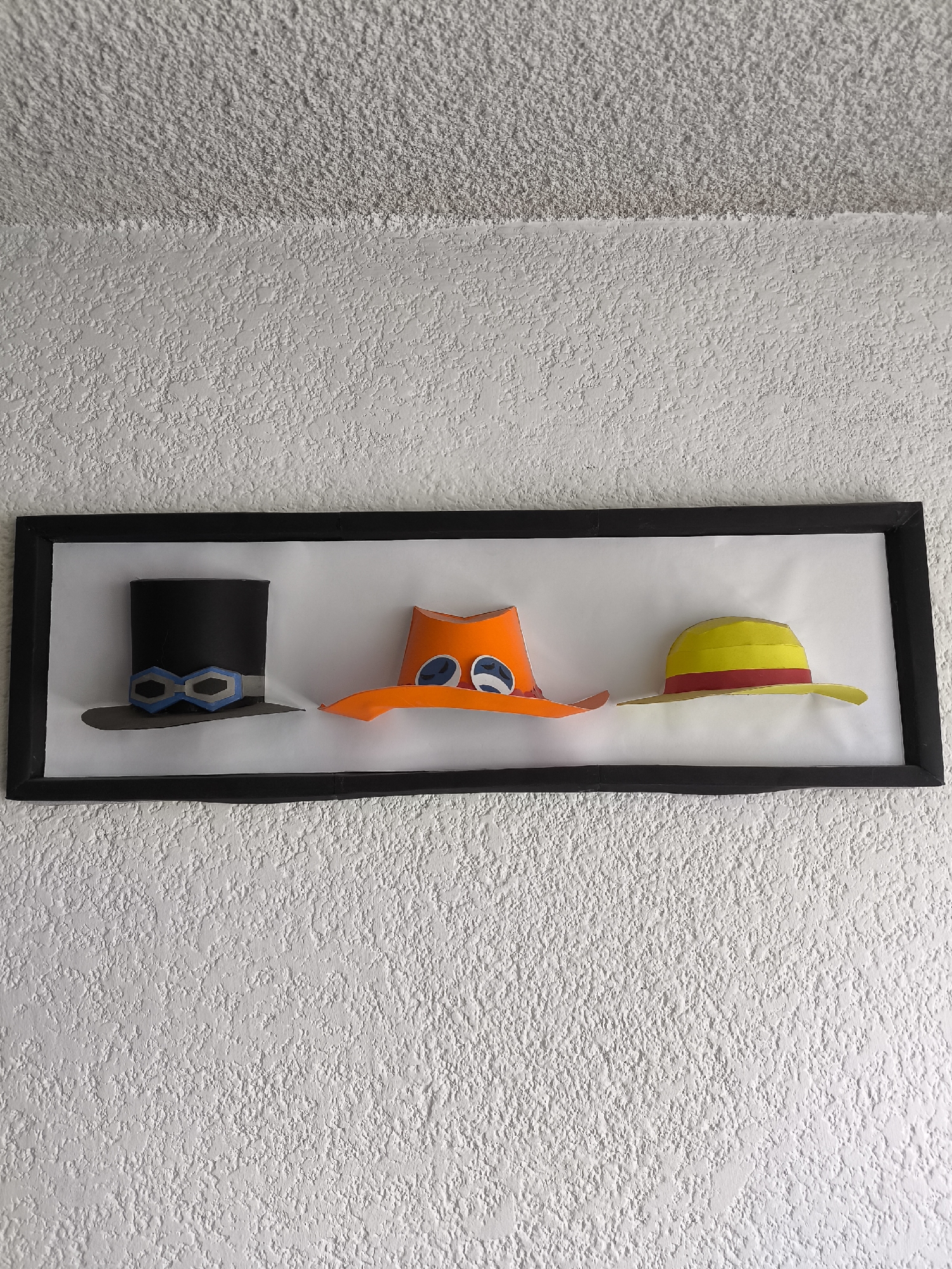 A framed artwork featuring three stylized hats is mounted on a textured white wall. The frame is black. From left to right, the hats are: a black top hat with blue and gray goggles, an orange hat with two large white eyes with blue pupils, and a yellow straw hat with a red band. The artwork is set against a white background. The wall itself has a rough, stucco-like texture. There are no people visible in the image. It is impossible to determine from this image if the photo was taken in Puebla, Mexico.