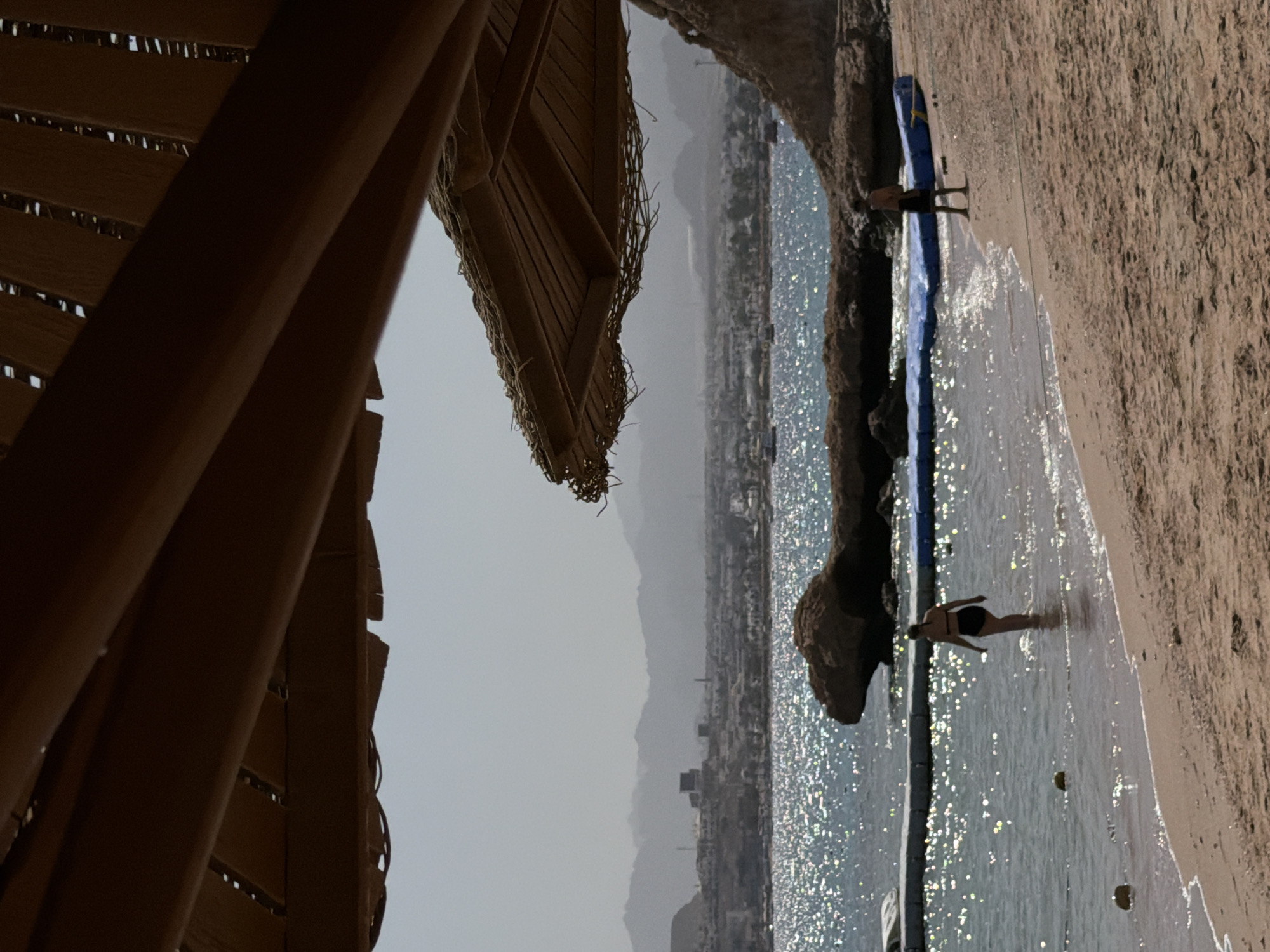 The image captures a coastal scene in Sharm El Sheikh, Egypt. The vantage point is partially obscured by a partially visible structure of what appears to be a thatched-roof hut. In the background, a coastline is visible, with buildings and hills stretching towards the horizon under a hazy sky. In the sea, there are dark rocks. Two people are seen in the water near the shoreline. The light on the water reflects brightly. The beach is visible along the coastline.