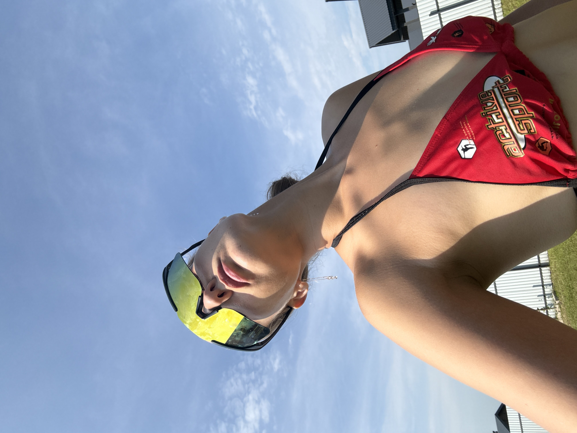 A low-angle medium shot captures a young woman in a red bikini and oversized yellow-green wraparound sunglasses, looking up at the sky. She has her head tilted back, her lips slightly parted, and a subtle expression on her face. A thin silver chain with a small charm dangles from her left ear. The background consists of a bright blue sky with wispy white clouds. In the distance, behind a white picket fence and some green grass, are parts of residential buildings with dark roofs. The scene appears to be set outdoors in a suburban area, possibly in Подшивалово, Russia, given the architectural style of the buildings. The lighting suggests it is a sunny day.