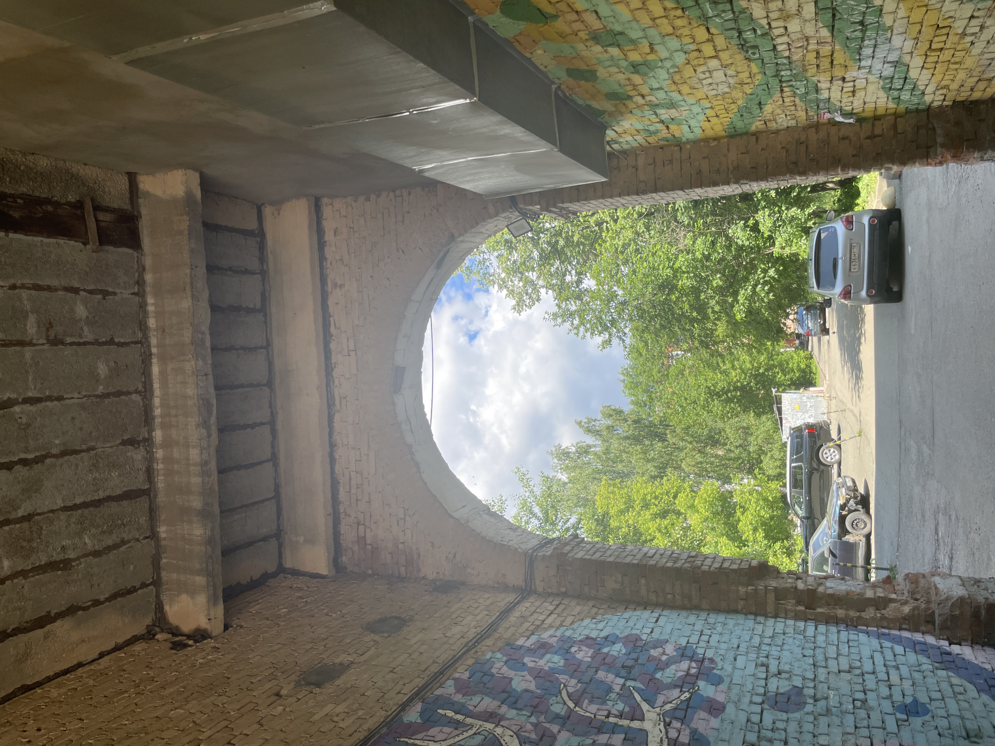 The image depicts an archway leading to an outdoor area with trees and parked cars. The archway is constructed of brick and concrete, with exposed concrete beams overhead. The walls of the archway are decorated with colorful murals. In the foreground, to the left, a brick pillar with a blue, purple, and white mosaic mural is visible. To the right, a brick pillar with a yellow and green striped mural is visible. Beyond the archway, several cars are parked on a paved surface, including a black SUV, a silver hatchback, and a dark-colored sedan in the distance. Lush green trees line the area behind the cars. The sky above is cloudy. The scene appears to be located in a residential or public outdoor space. The license plate on the silver hatchback reads "K636MT 18," indicating the vehicle is registered in Russia. Based on this registration detail, the location can be identified as Izhevsk, Russia.