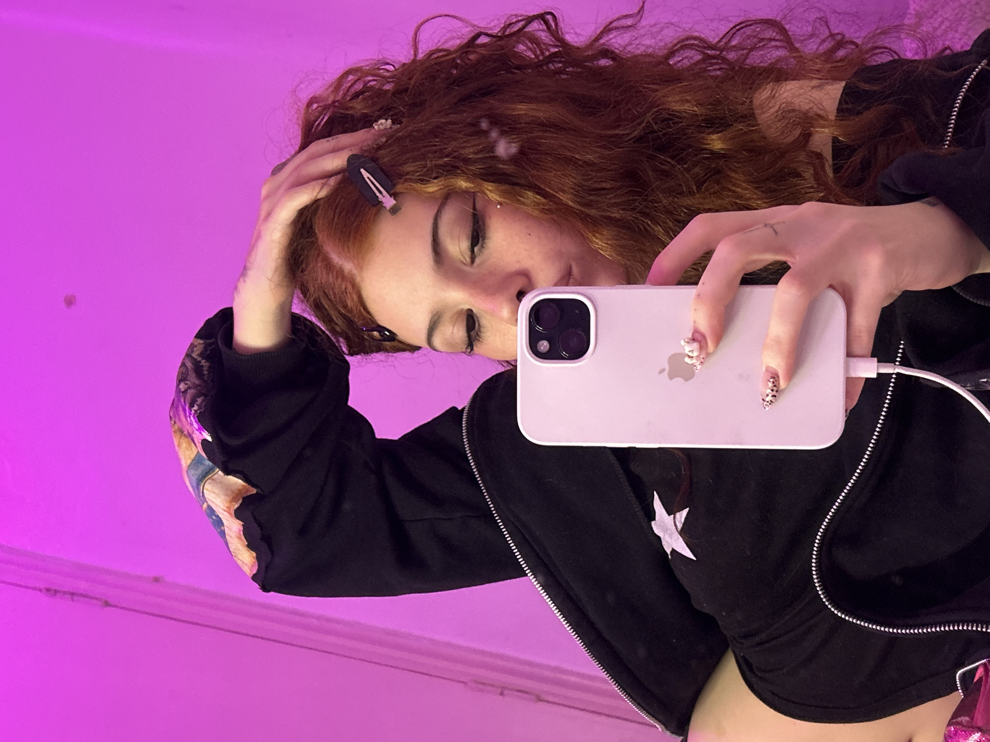A woman with auburn curly hair is taking a selfie in what appears to be an indoor location with pink lighting. She is wearing a black zip-up jacket with a star design. Her phone, possibly an iPhone, is white and is plugged into a charger. Her nails are decorated with a patterned design. A black hair clip is visible in her hair. The setting is indistinct and there is no indication of Buenos Aires, Argentina.