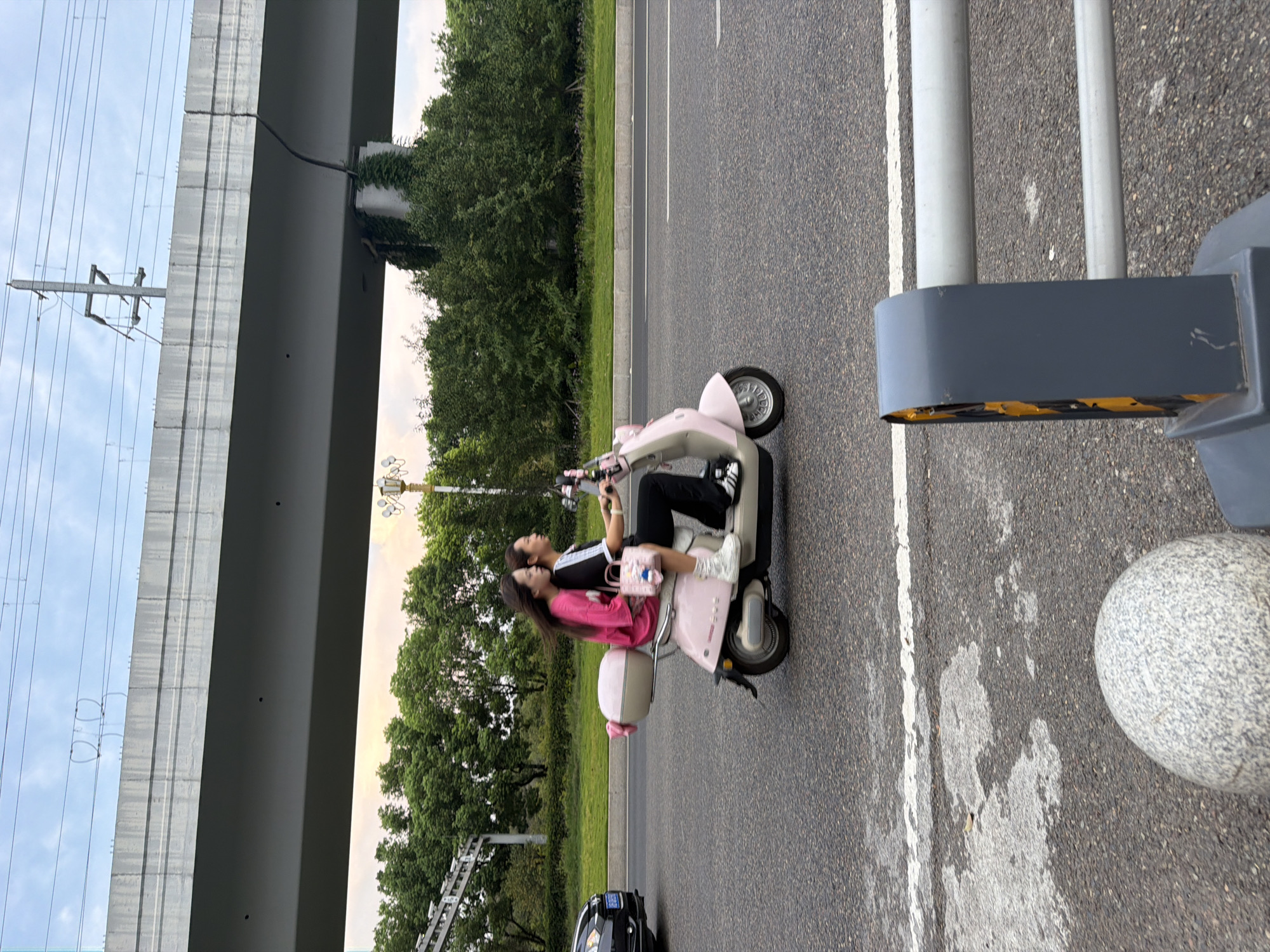 Two people, a male driver and a female passenger, are riding a pink scooter on a paved road. The man is wearing a black shirt with white stripes and black pants, while the woman is wearing a bright pink top and white sneakers. They are riding side-by-side on the scooter, with the woman holding a small pink bag. The scooter has a pink storage compartment on the back with a pink bow attached. In the background, there is a large concrete overpass, trees, and a cloudy sky. A black car is visible in the distance on the left side of the frame, and a gray bollard with reflective yellow strips is in the foreground on the right. The image appears to have been taken in Jiangjin District, Chongqing, China, given the context of the request.