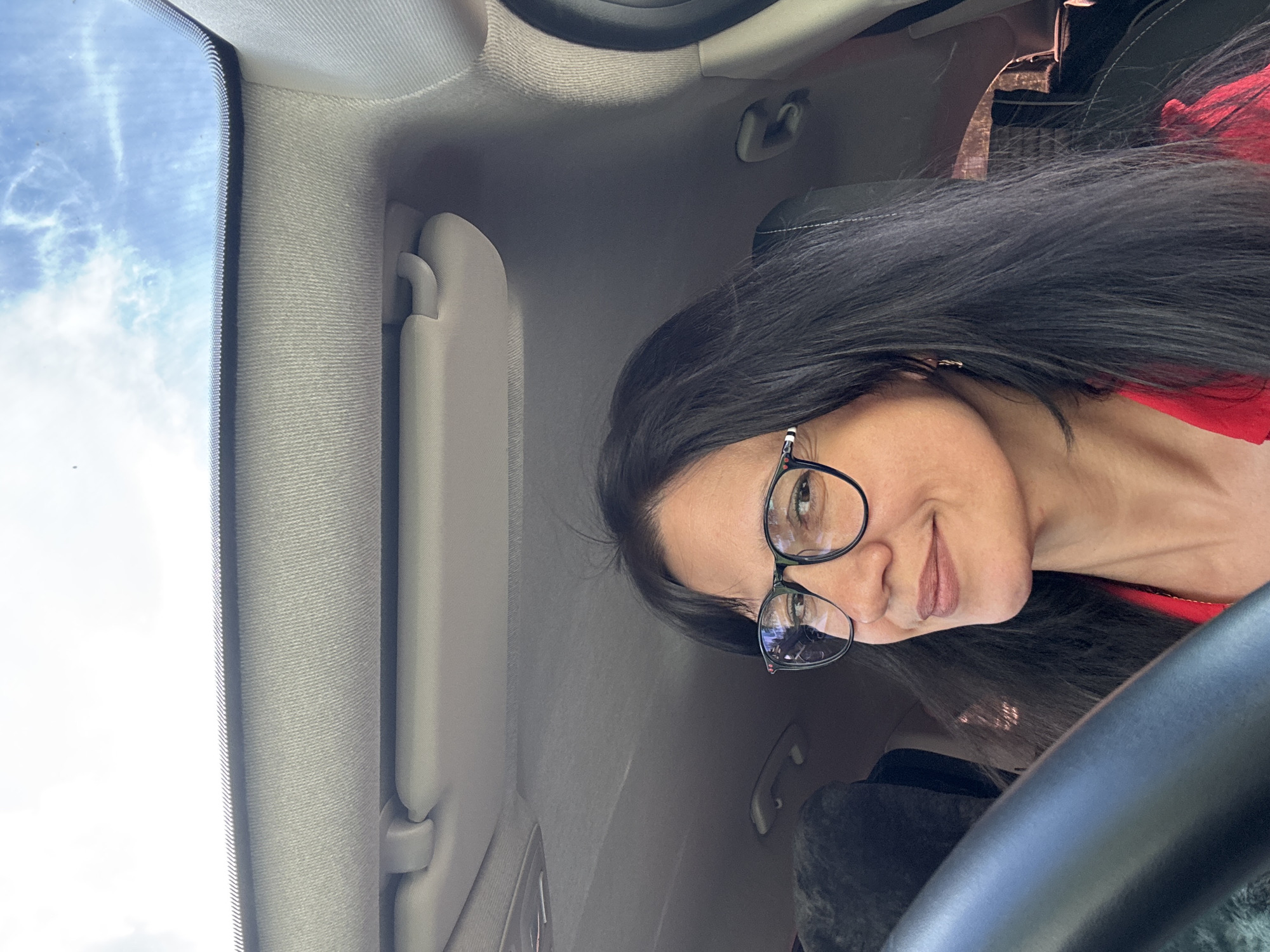 The image shows a woman with long, dark hair and glasses smiling as she sits in the driver's seat of a car. She is wearing a red top. The interior of the car is visible, including the steering wheel, dashboard, and a sun visor. The sky is visible through the windshield, with some clouds. The image is a selfie taken from the perspective of the driver. The location is not specified in the image or its metadata.
