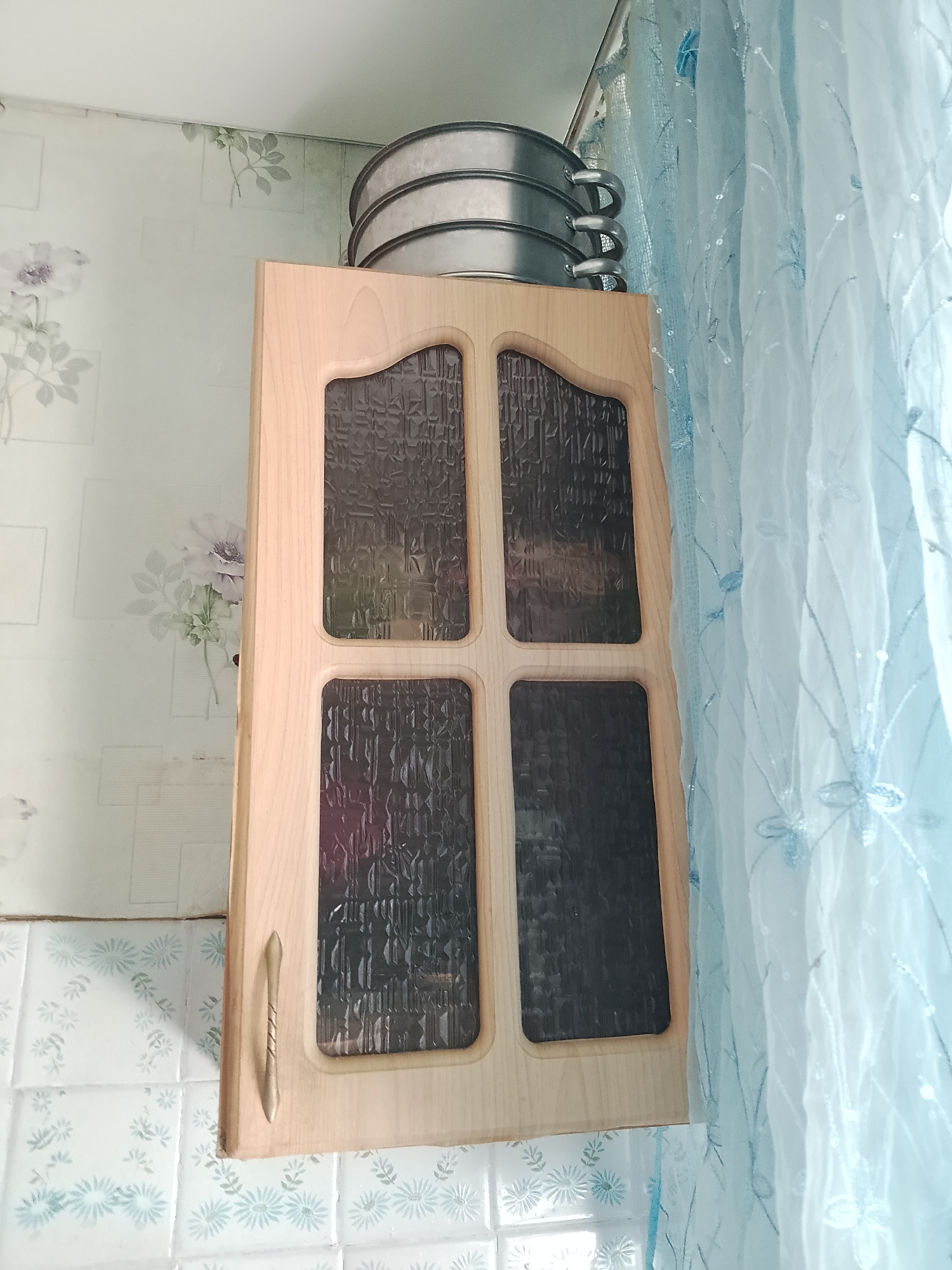 The image depicts a close-up view of a kitchen cabinet with a wooden frame and four textured glass panels. A silver cabinet handle is attached to the left side of the cabinet door. Above the cabinet, a stack of three silver cake pans is visible. To the right of the cabinet, a sheer, light blue curtain with an embroidered floral pattern hangs. The background shows floral patterned wallpaper. The scene is likely an interior shot, possibly in a residential kitchen, in the city of Abay, Kazakhstan.