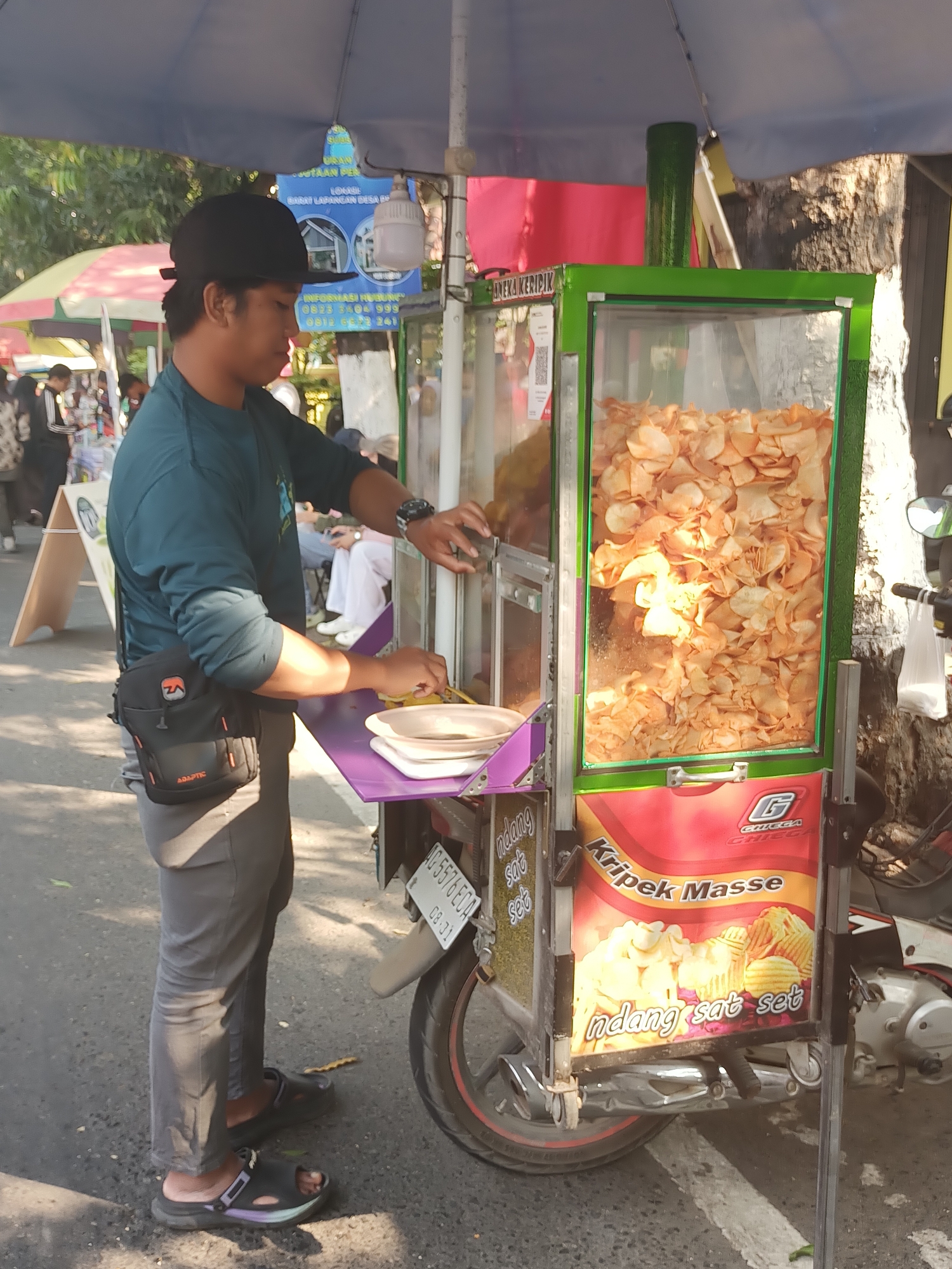 A young man wearing a black baseball cap, a dark teal long-sleeved shirt, grey pants, and black sandals stands at a food cart in the city of Pare, Indonesia. He has a black cross-body bag with an orange and white logo on his left hip. The man is actively engaged with the food cart, holding a tool in his right hand and looking towards the cart. The food cart itself is green and glass, filled with a large quantity of thinly sliced, golden-brown potato chips. The cart has various labels and graphics, including one that reads "Kripek Masse" and another with "G CHIEGA". A motorcycle is parked to the right of the cart, partially visible. The scene takes place outdoors on a paved street, with a tree and a blue umbrella providing shade. In the background, other people and food stalls are present, suggesting a market or street vendor area.