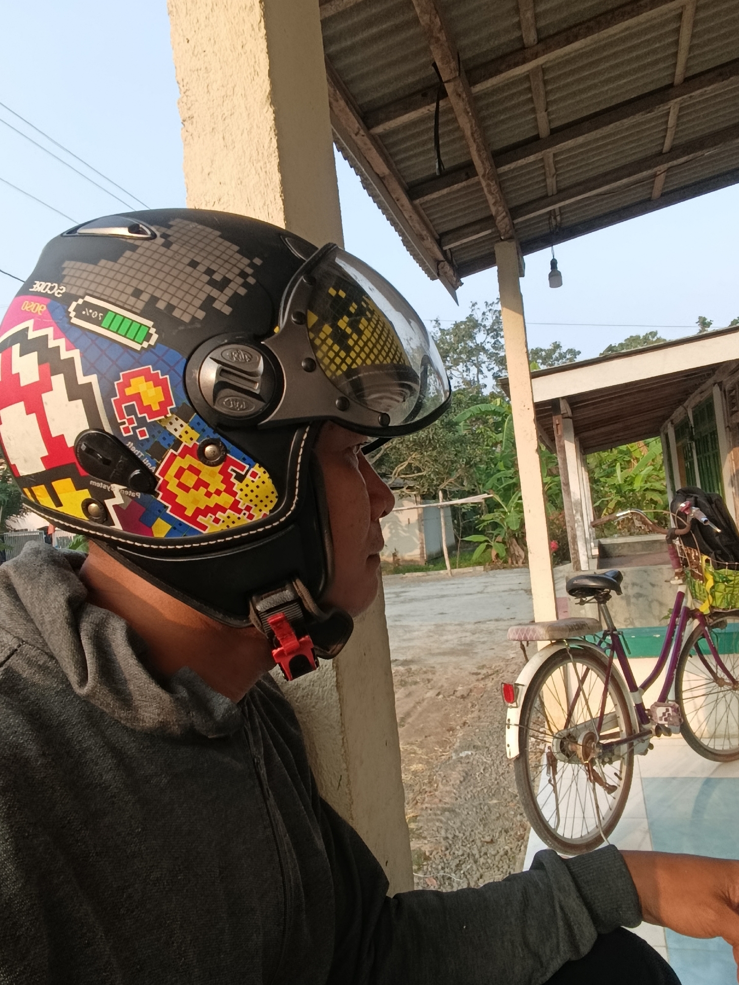 A person wearing a black helmet with colorful pixelated graphics is pictured in Sarwadadi, Indonesia. The helmet features designs resembling a battery charge indicator and video game sprites. The person is wearing a grey hooded sweatshirt. In the background, a purple bicycle with a basket is parked. The scene is set outdoors, under a corrugated metal roof supported by wooden beams. Trees and vegetation are visible behind the structure, and power lines stretch across the sky. The lighting suggests it is daytime.