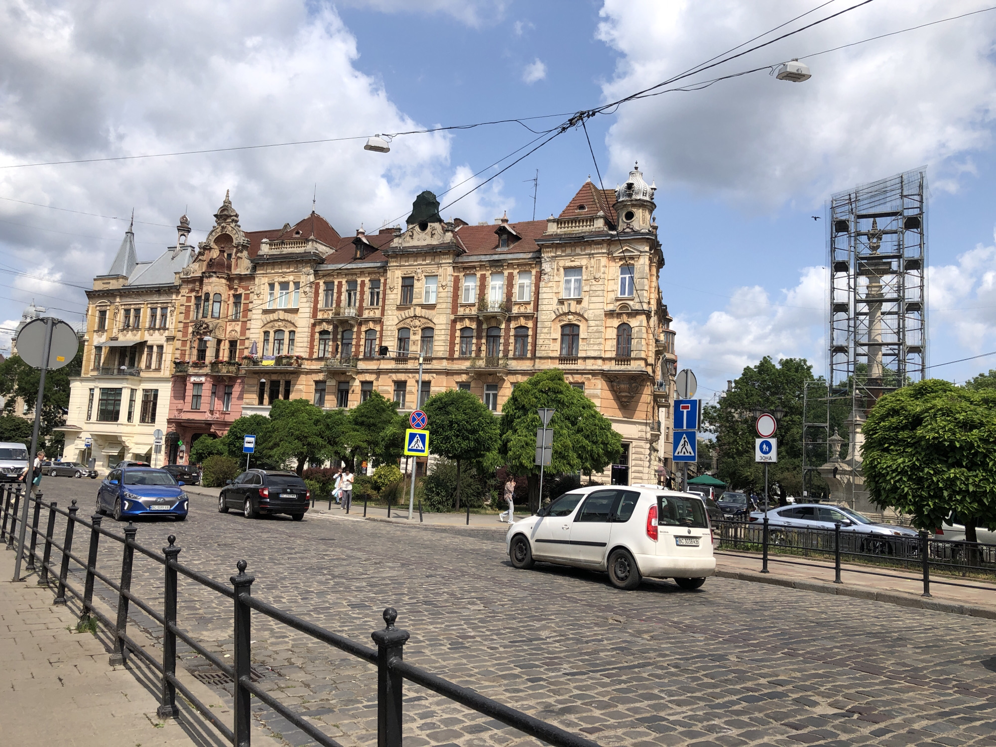 The image captures a view of a city street in Lviv, Ukraine, under a partly cloudy sky. In the foreground, a cobblestone street is bordered by a black metal fence. Several cars are visible on the street: a blue sedan, a black station wagon, and a white hatchback. Pedestrians are walking on the sidewalk. Behind the street, a large, ornate building with a yellow facade and a red tile roof dominates the scene. To the right of this building, a tall structure covered in scaffolding stands against the sky. Trees and other smaller buildings are also present, contributing to the urban landscape. Traffic signs and overhead wires are scattered throughout the scene. The overall impression is of a bustling city street with historical architecture and modern elements.