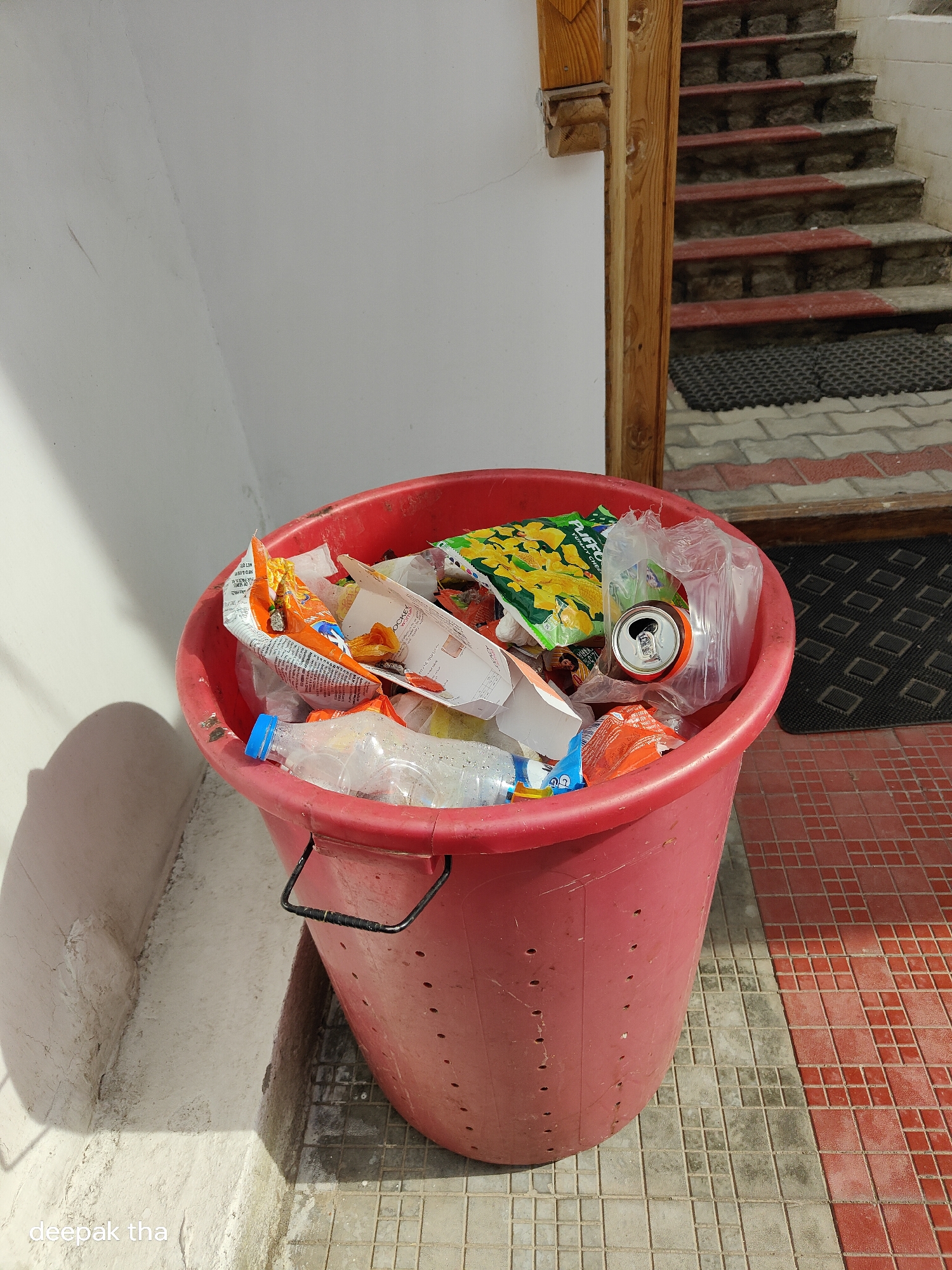 A large, red plastic trash can is overflowing with assorted waste, including plastic bottles, snack wrappers, and a beverage can. The trash can is placed on a tiled outdoor surface, with a white wall and a wooden staircase visible in the background. The environment suggests an urban setting, possibly in Leh, India, given the architecture and paving style. The lighting indicates it is daytime, with shadows cast by the sun. The scene is static, with no people or animals present.