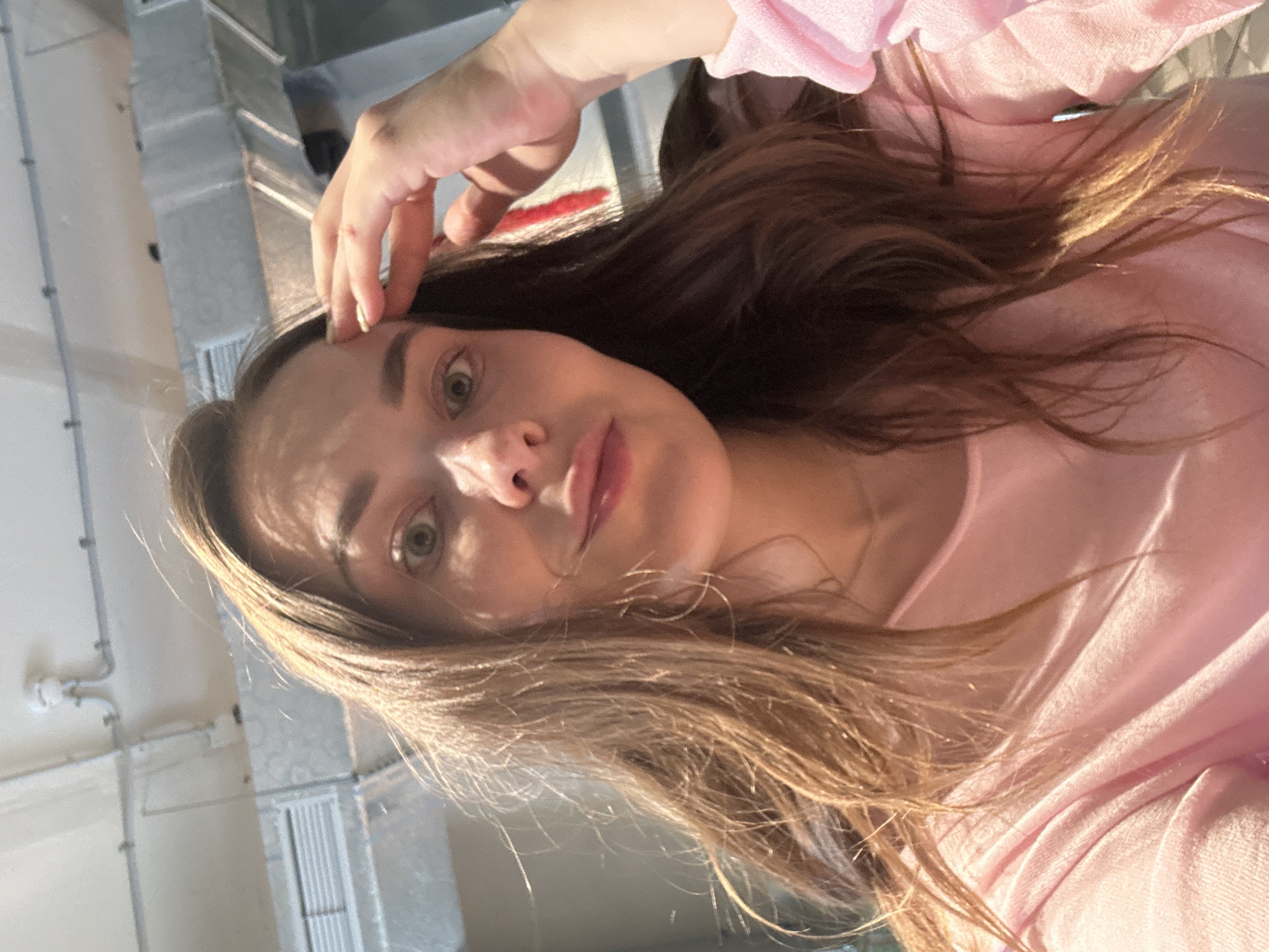 The image is a close-up, low-angle selfie of a young woman with long, light brown hair. She is wearing a pink top. Her hand is raised to her forehead, with her fingers gently touching her hair. Sunlight is casting shadows across her face and hair, creating a warm and ethereal effect. Her eyes are open and looking towards the camera, and her lips are slightly parted. The background appears to be an indoor setting with industrial elements, such as exposed pipes and ventilation systems, suggesting a warehouse or studio environment. The overall mood of the image is relaxed and contemplative, with the soft lighting and the woman's gentle gesture contributing to a sense of introspection. The time of day is likely daytime, given the strong sunlight. There is no visible text in the image. The location context is Perm, Russia.