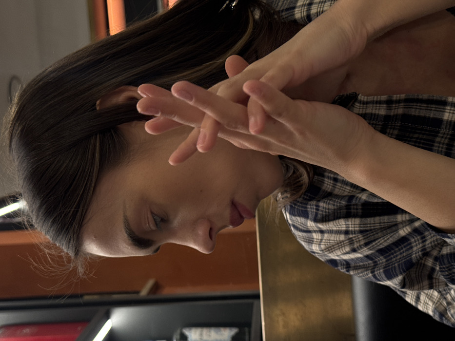 The image presents a close-up, mid-profile view of a female individual within an indoor commercial establishment, specifically in Verona, Italy. The subject, oriented towards the left, has long, dark brown hair and fair skin. Her facial features include pronounced eyebrows, a straight nose, and full lips, with her gaze directed downward. She is wearing a blue and white plaid button-down shirt, unbuttoned at the collar. Her hands are clasped together in front of her upper chest, with fingers interlocked, displaying light-colored nail polish. The background, out of focus, includes a dark seating surface in the lower left, a horizontal golden-toned barrier, and beyond that, an orange-hued wall with vertical strip lighting. A clear glass bottle with a white and blue label is visible on a shelf to the far left.