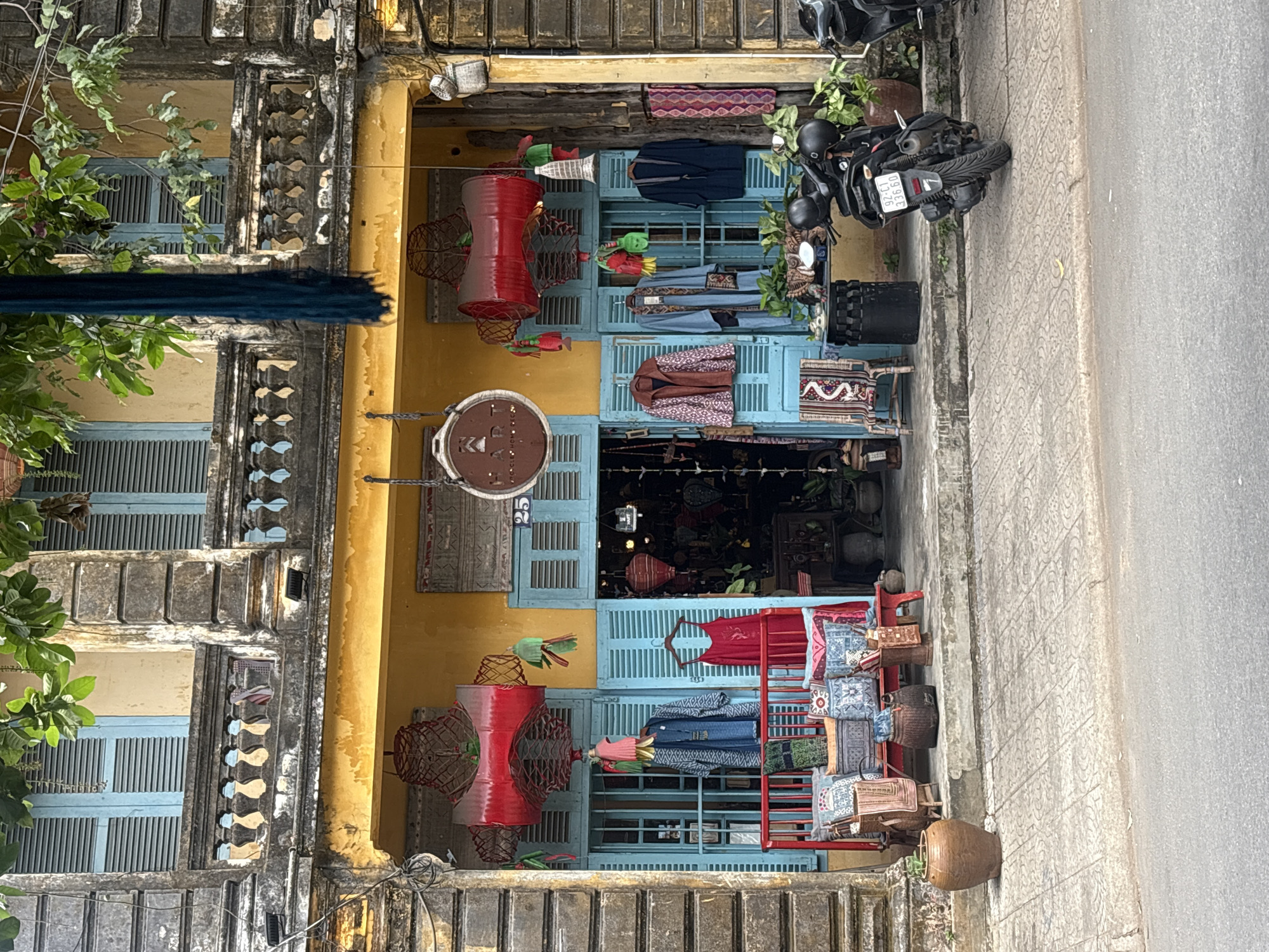 This is a medium shot of a shop in Hoi An, Vietnam, featuring traditional Vietnamese architecture and goods. The shop's facade is painted a vibrant yellow, contrasting with the weathered stone of the building's upper story. Blue louvered shutters frame the shop entrance, which displays a variety of items for sale.
In the foreground, a patterned tiled walkway leads to the shop. Two black scooters are parked on the right side of the image, with one facing the viewer. A large, dark brown ceramic pot sits on the left side of the walkway.
The shop itself has a red railing in front of the entrance, adorned with several patterned cushions and woven baskets. Behind the railing, clothing items hang from hooks, including a red dress and a patterned jacket. Large red cylindrical lanterns hang above the entrance, alongside decorative woven baskets and green and red tassels.
The interior of the shop is visible through the blue shutters, revealing more clothing, lanterns, and decorative items. A sign with the number "25" is visible above the right shutter. The overall impression is of a bustling market stall filled with colorful textiles and traditional crafts.