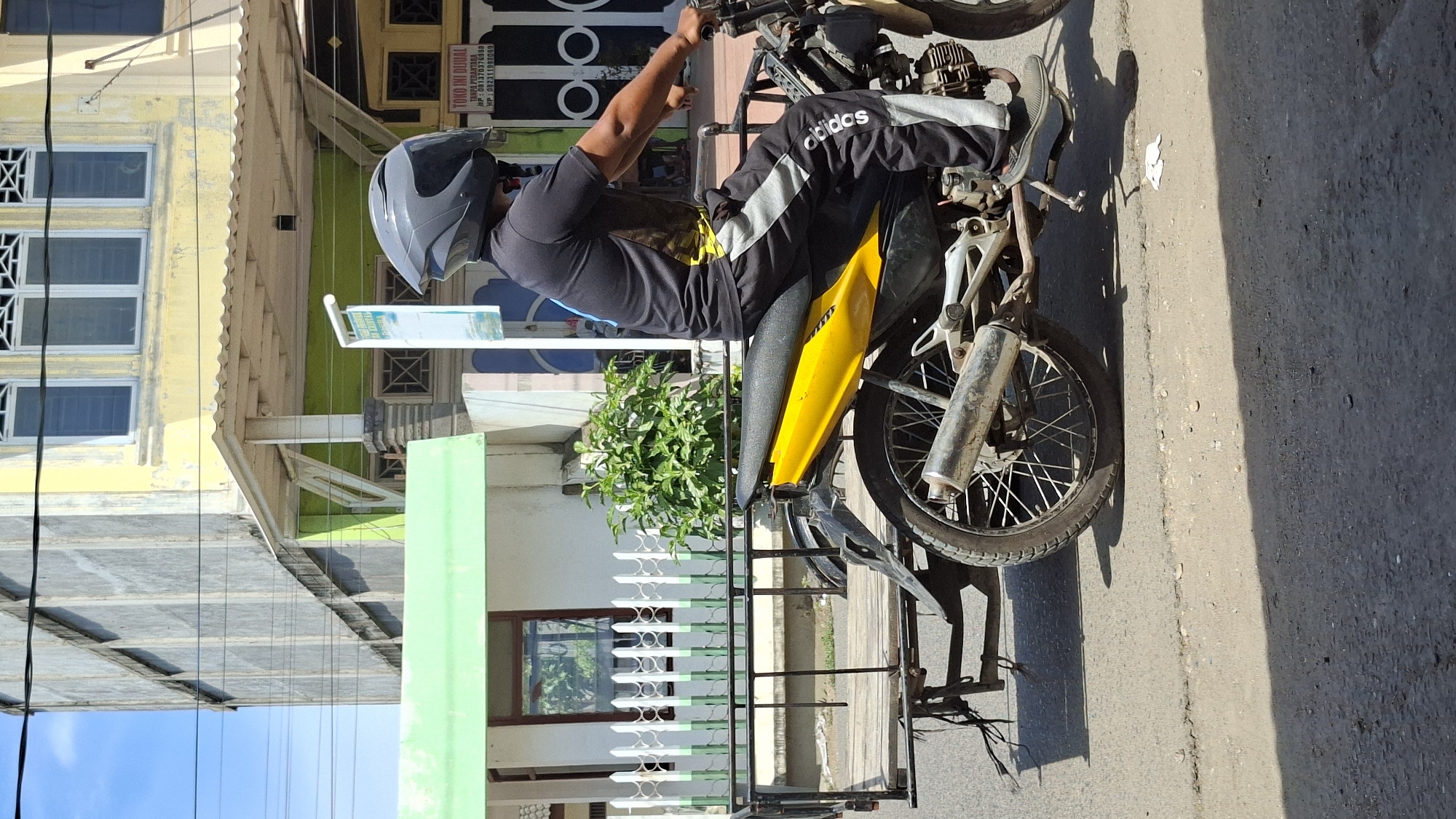 A person wearing a black t-shirt, black pants with grey stripes, and a grey helmet is riding a yellow motorcycle with a sidecar. The motorcycle has a silver exhaust pipe and spoked wheels. The sidecar is made of wood and metal bars. The scene takes place on a paved road in front of buildings with yellow and green facades. A white picket fence and green bushes are visible behind the motorcycle. A sign with Indonesian text is also present. The weather appears to be sunny with shadows cast on the road. The image was taken in Lhokseumawe, Indonesia.
