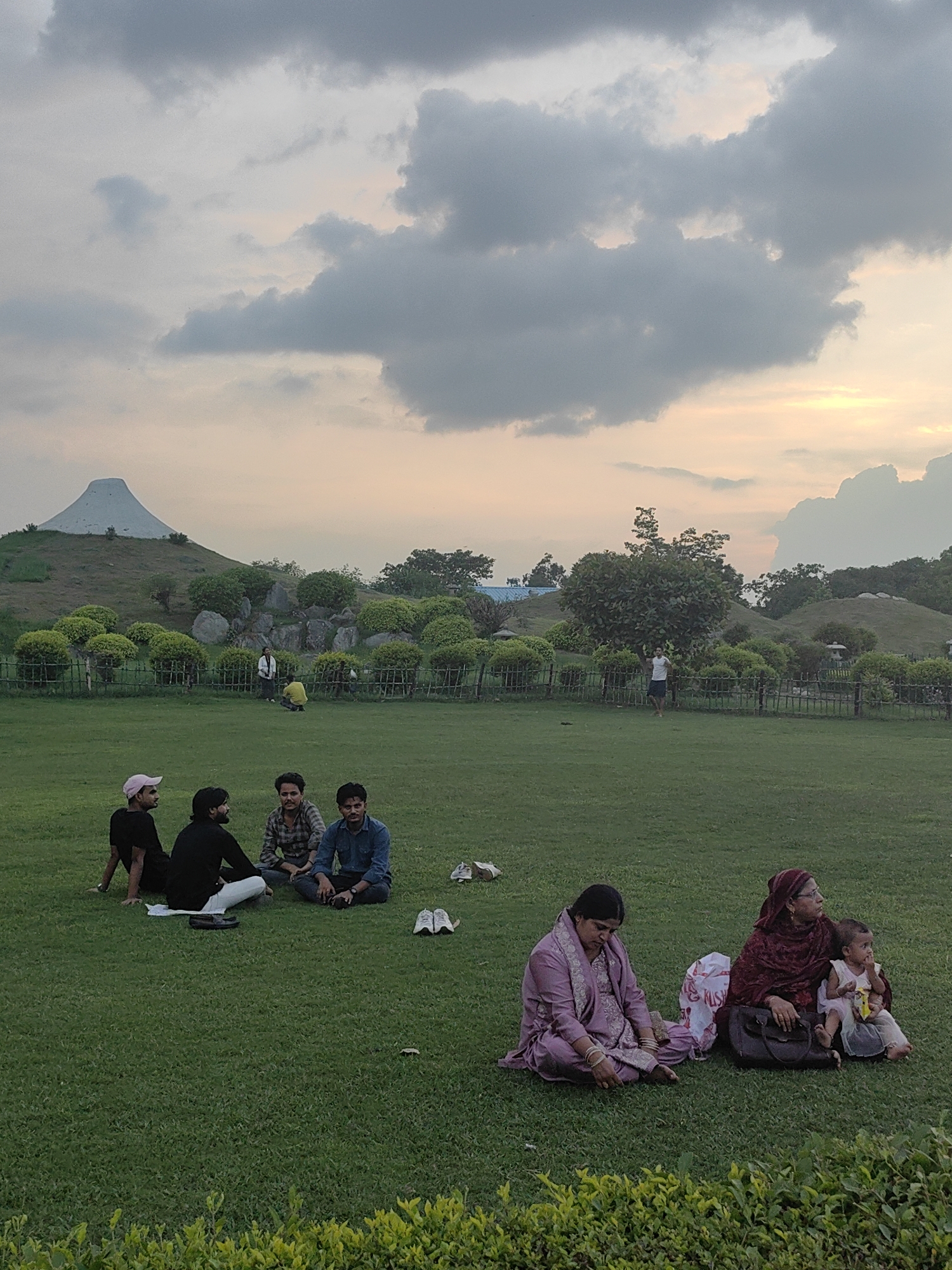 In Delhi, India, a group of people are enjoying a day at a park. The image captures a serene atmosphere with a vast expanse of green lawn, dotted with trees and manicured bushes. In the background, a striking conical structure, possibly an art installation or a monument, rises against a sky filled with dramatic clouds, tinged with the soft hues of sunset. A group of four young men are seated on the grass in the foreground, engaged in conversation. Further on, two women and a child are also resting on the lawn, with one woman holding the child. The park appears to be a popular spot for leisure, with various individuals and small groups scattered across the open spaces, enjoying the pleasant weather and the picturesque surroundings. The overall impression is one of a peaceful, communal gathering in an urban green space.