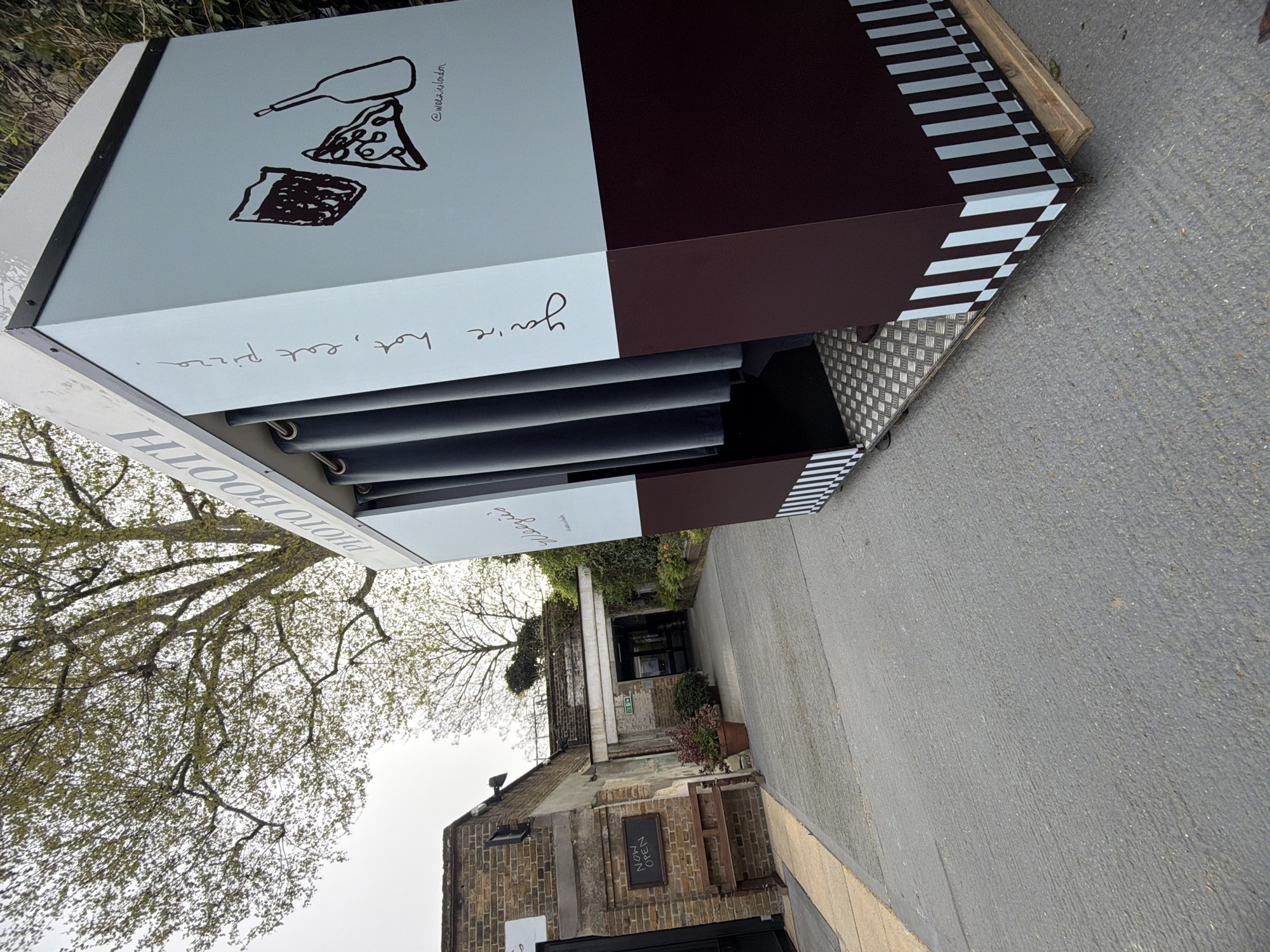 A photo booth sits on the pavement next to a building, with the brand name 'Mojeez' written on the booth. The booth is painted white and dark brown, with a pizza drawing and the words, "You're hot, eat pizza." A tree is on the left of the photo. In the background is a building with a sign that says, "NOW OPEN." The location is in the city of London, United Kingdom.