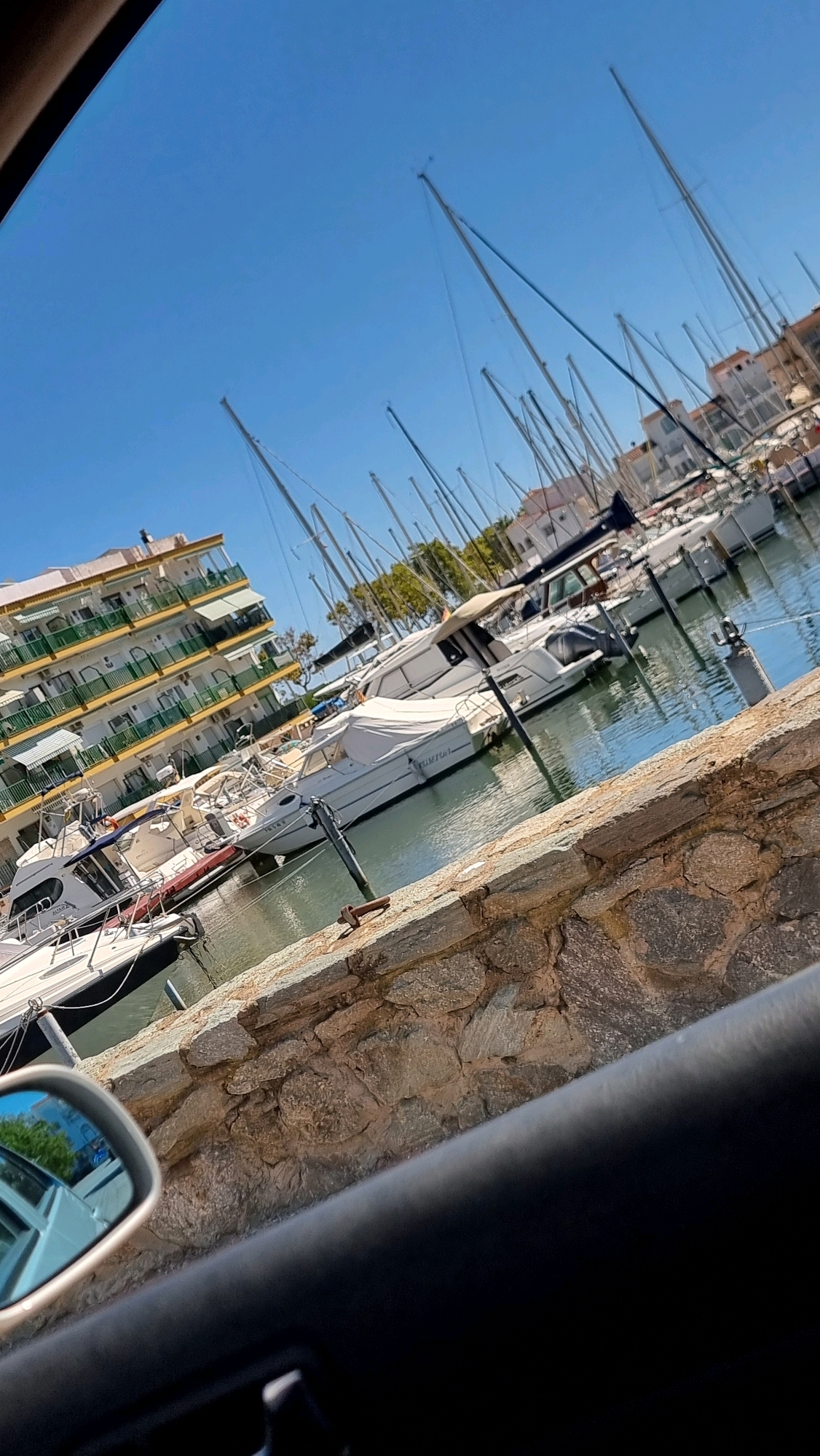 The image depicts a marina in the city of Castelló d'Empúries, Spain, on a clear, sunny day. Several sailboats and motorboats are docked, with masts prominently visible against the bright blue sky. In the background, a multi-story building with a yellow facade and green balconies lines the waterfront. A stone wall runs along the foreground, partially obscuring the view of the marina and the reflected sky in a car's side mirror. The water in the marina is calm, with reflections of the boats and the sky. No people are visible in the scene.