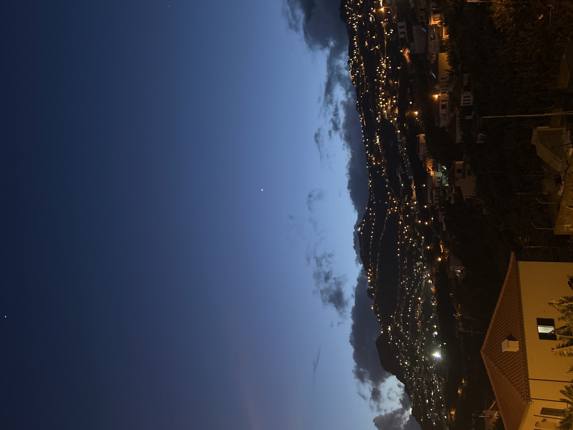The image presents an evening view of Câmara de Lobos, Portugal. A dark blue sky dominates the scene, punctuated by a few visible stars. To the right, a steep hillside is densely populated with buildings, their lights creating a visually prominent network along the slopes. A building with a terracotta roof and lit windows is visible in the foreground. Clouds are interspersed along the hillside and in the sky, adding texture to the composition.