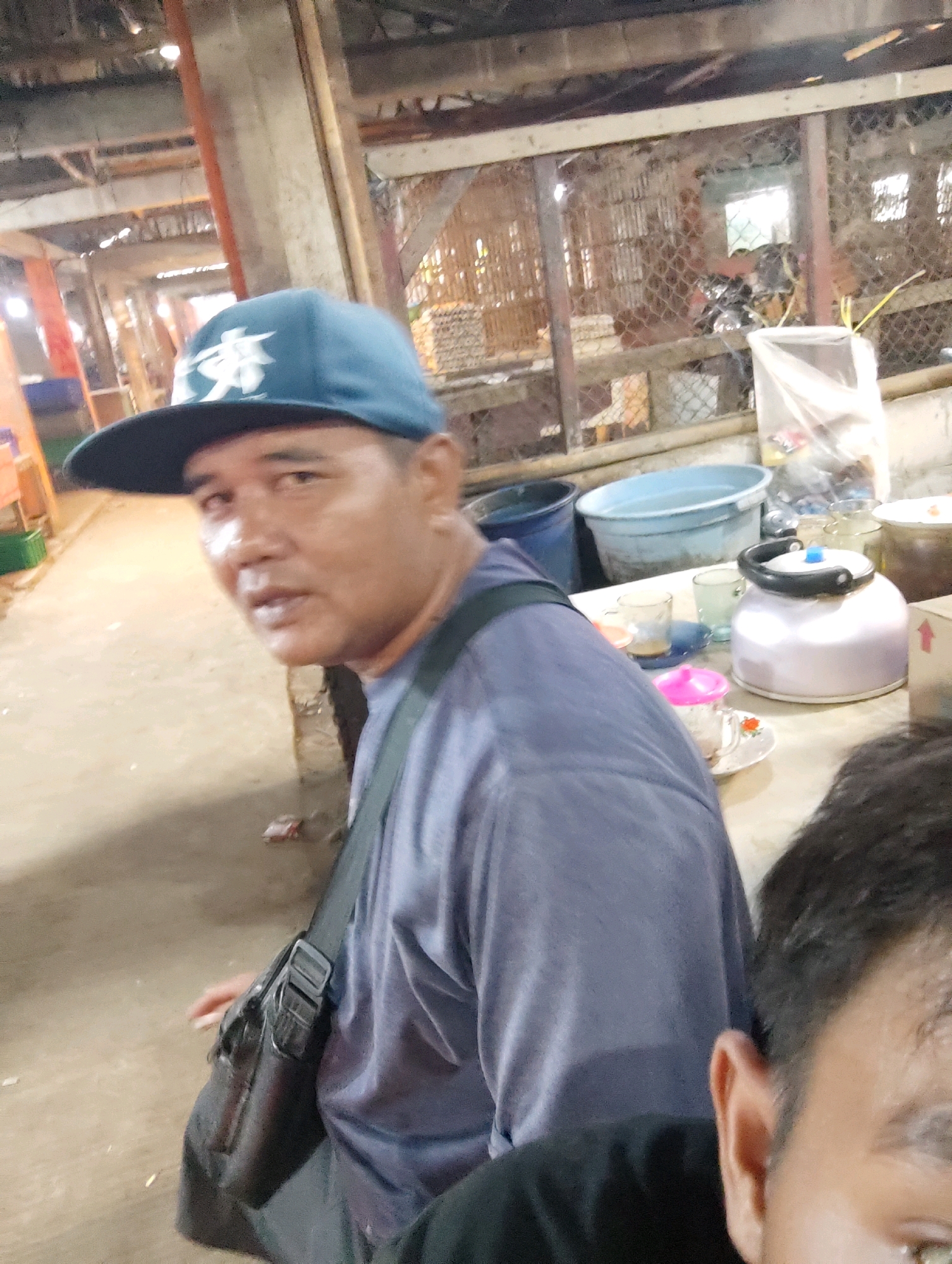 A man wearing a blue baseball cap with a white emblem and a purple t-shirt is in the foreground. He has a black messenger bag slung across his chest. In the background, there are cages that appear to house chickens, and tables with various items including buckets, a kettle, and glasses. The scene appears to be an outdoor market or a similar establishment in Karawang, Indonesia, given the context provided.
