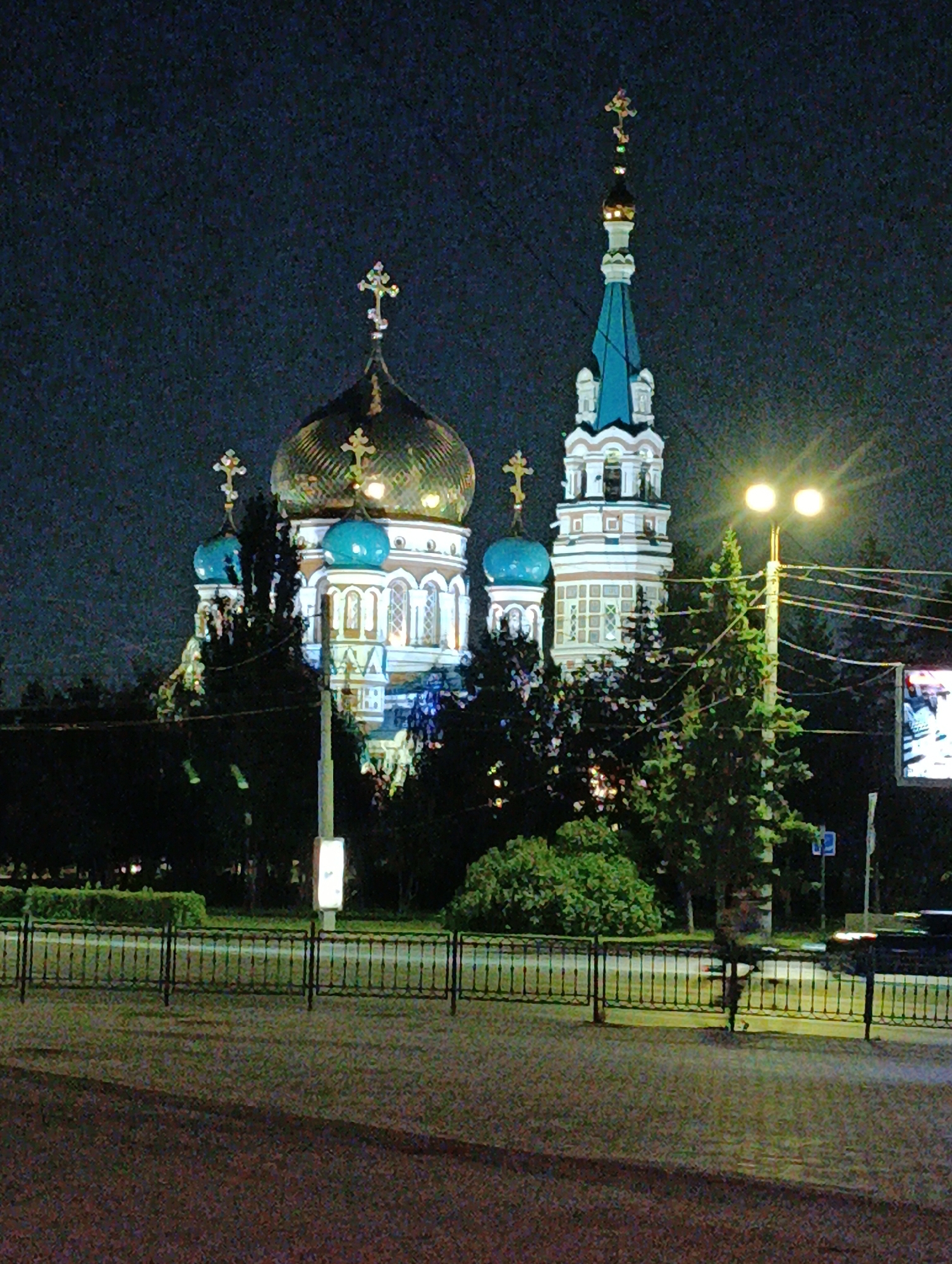 The image shows the Assumption Cathedral in Omsk, Russia at night. The cathedral is an Orthodox church with a large golden dome and a tall, slender bell tower topped with a blue spire. It is illuminated by lights, creating a warm glow. In the foreground, there is a black metal fence, a sidewalk, and some trees. A street light with two lamps is visible to the right of the cathedral, and power lines are strung across the scene. A car is partially visible on the right side of the image, and a billboard is also present. The sky is dark, indicating nighttime.